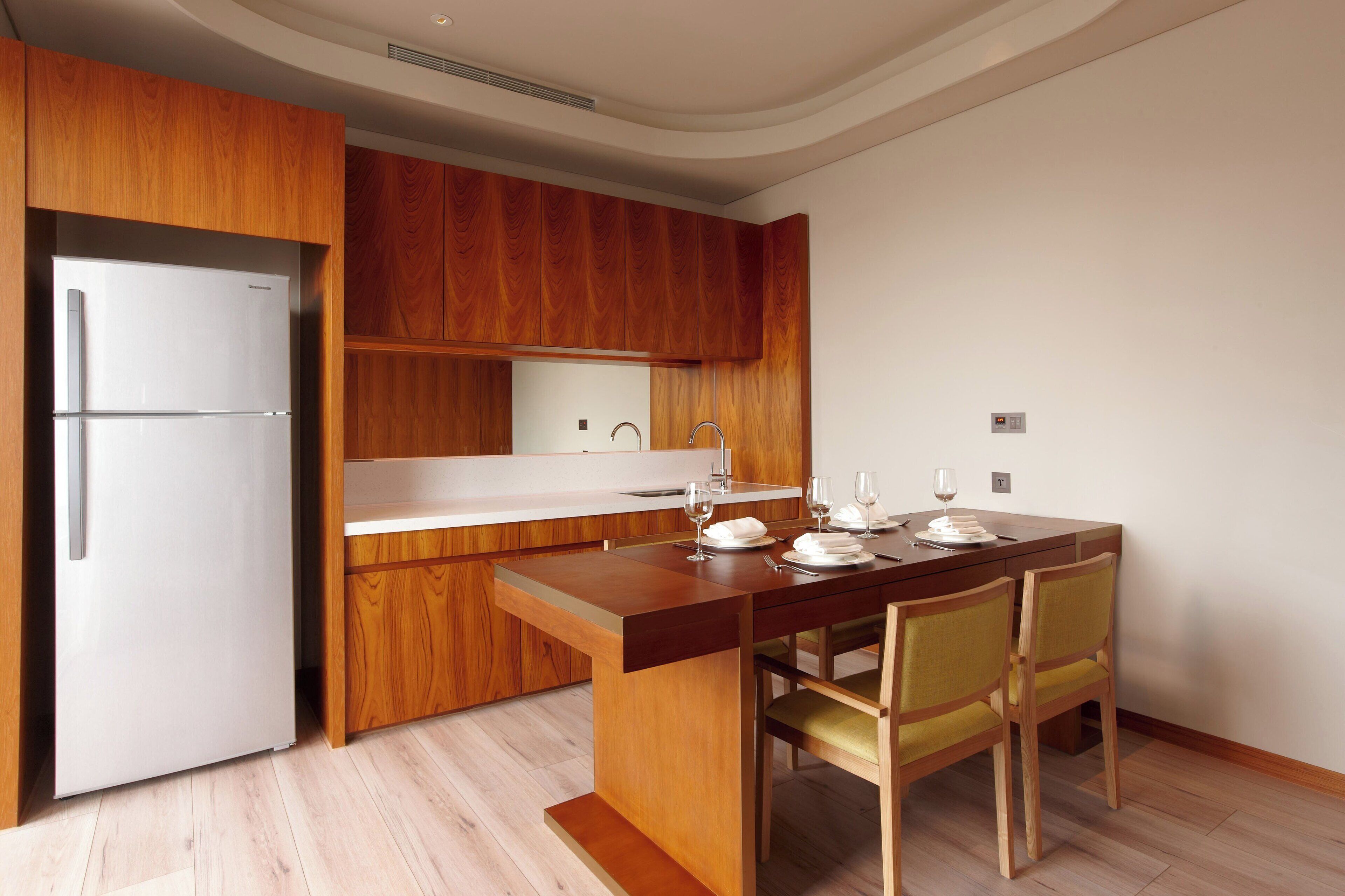 A modern kitchen with wooden cabinets, a white refrigerator, a wooden dining table with chairs, and a white countertop.