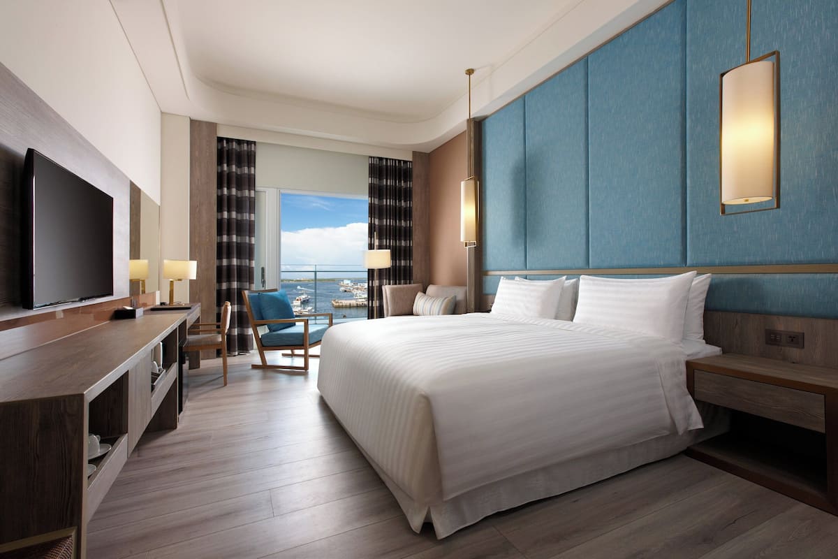 Habitación de hotel con una cama grande, televisión, escritorio con silla y vista al mar.