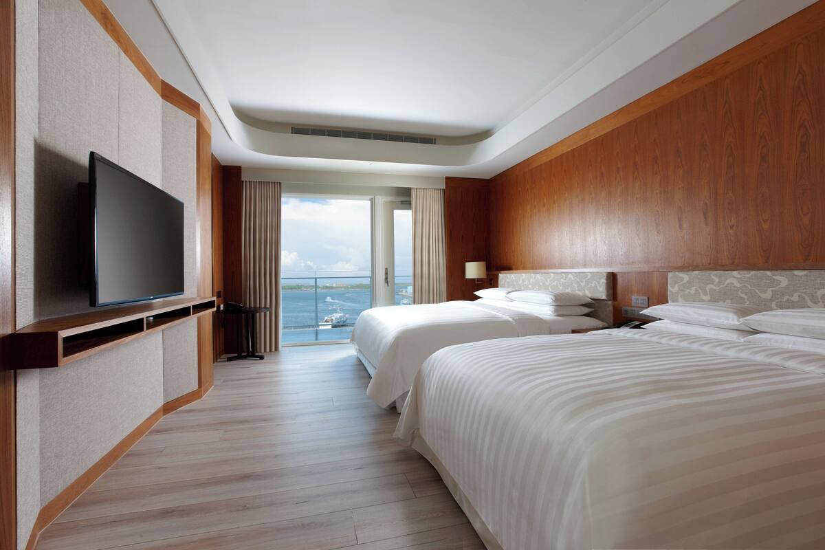 Habitación de hotel con televisor grande, dos camas, cabecero de madera y vista al mar.