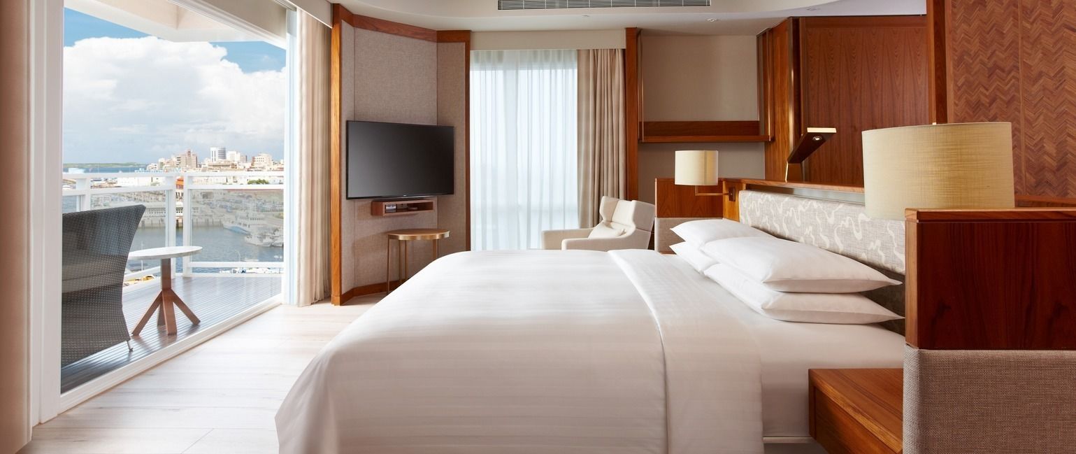Quarto de hotel com uma cama grande, televisão, poltrona, escrivaninha e vista para a cidade.