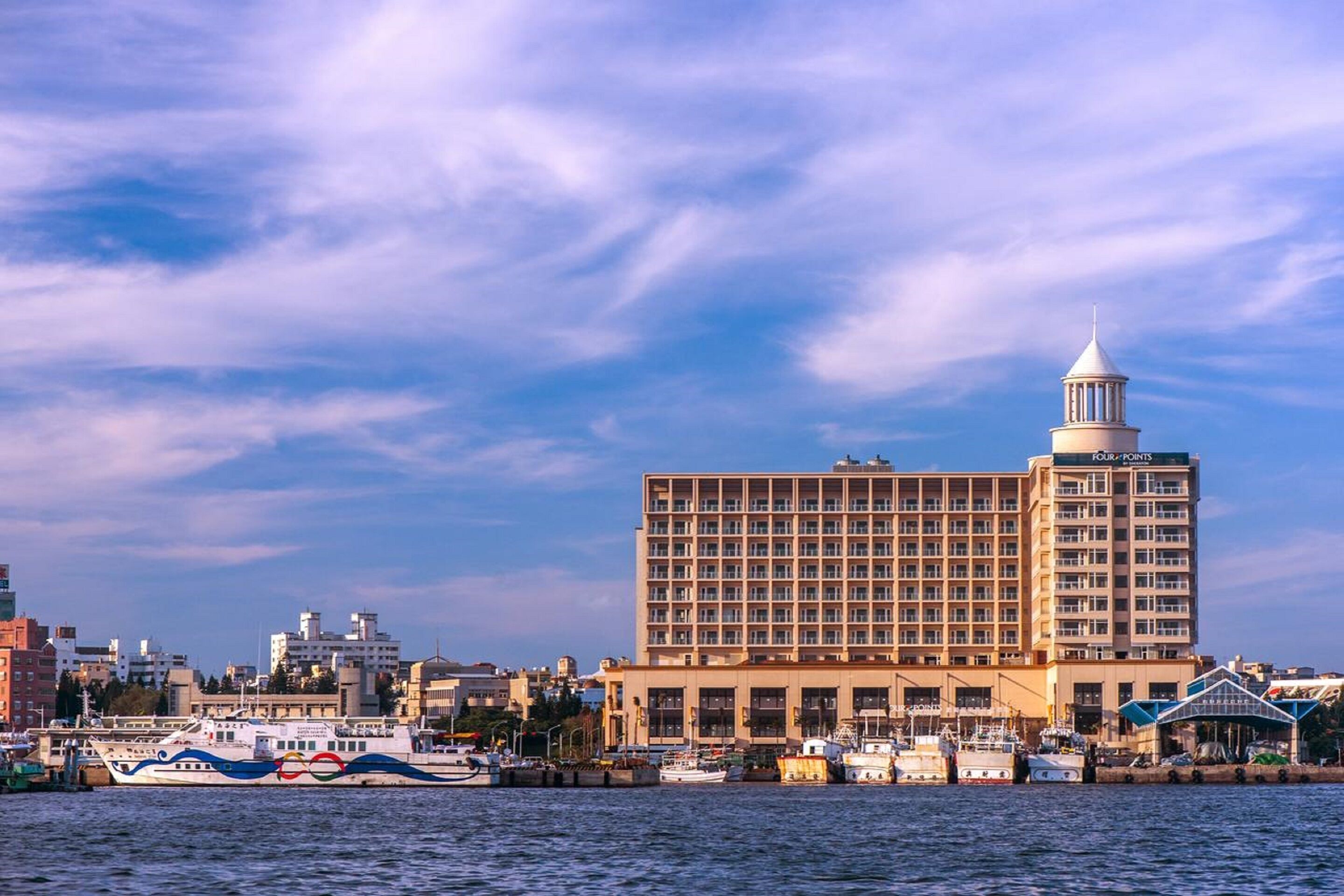 A large hotel with a prominent tower, located near a waterfront with boats docked.