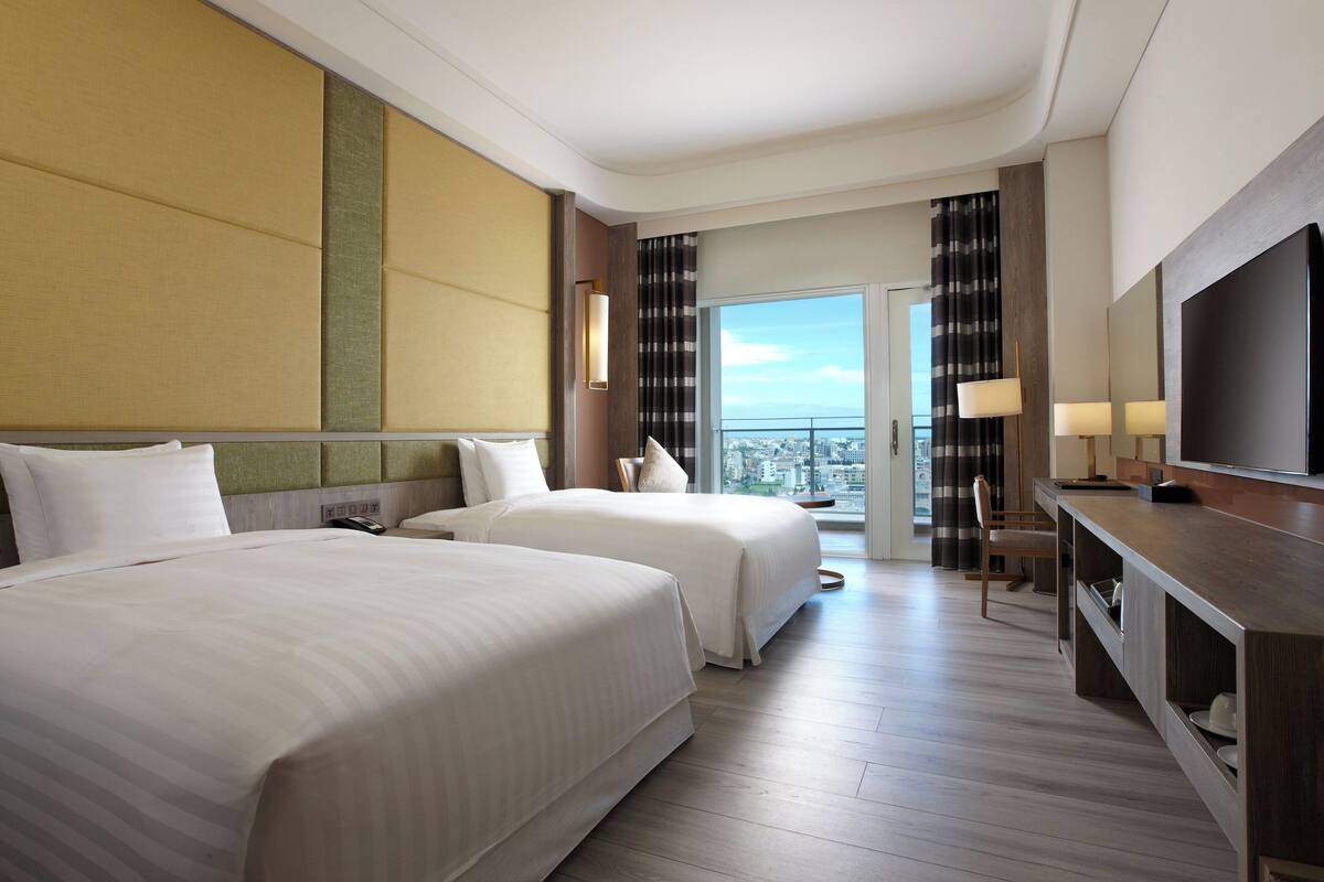 Habitación de hotel con dos camas, un escritorio, un televisor y un balcón con vista a la ciudad.