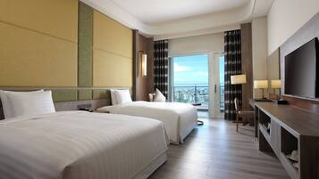 A hotel room with two beds, a desk, a TV, and a balcony with a city view.