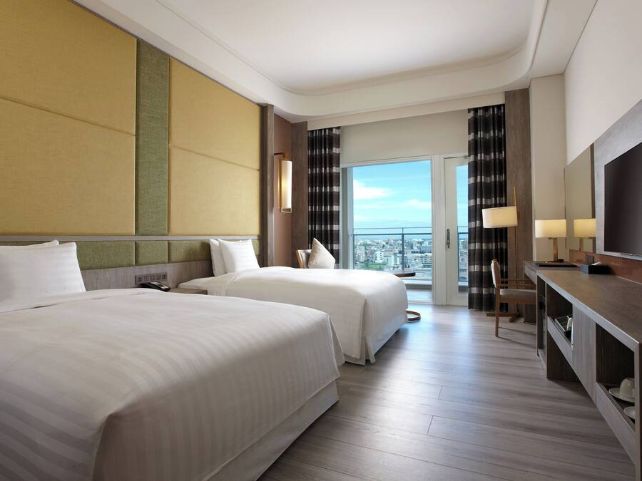 A hotel room with two beds, a desk, a TV, and a balcony with a city view.