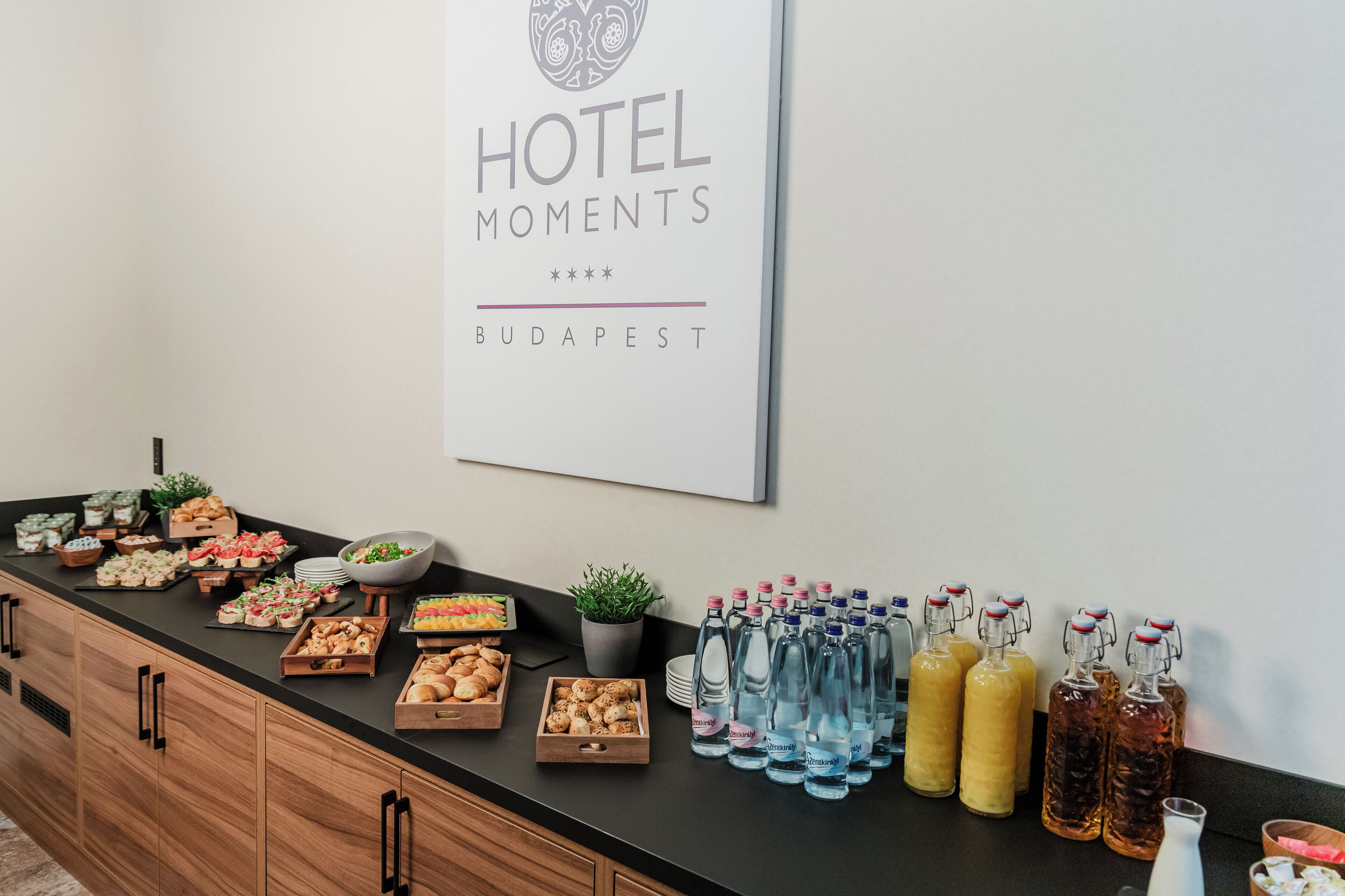 A buffet setup with various food items and beverages, a sign indicating 'HOTEL MOMENTS BUDAPEST', and wooden cabinets.