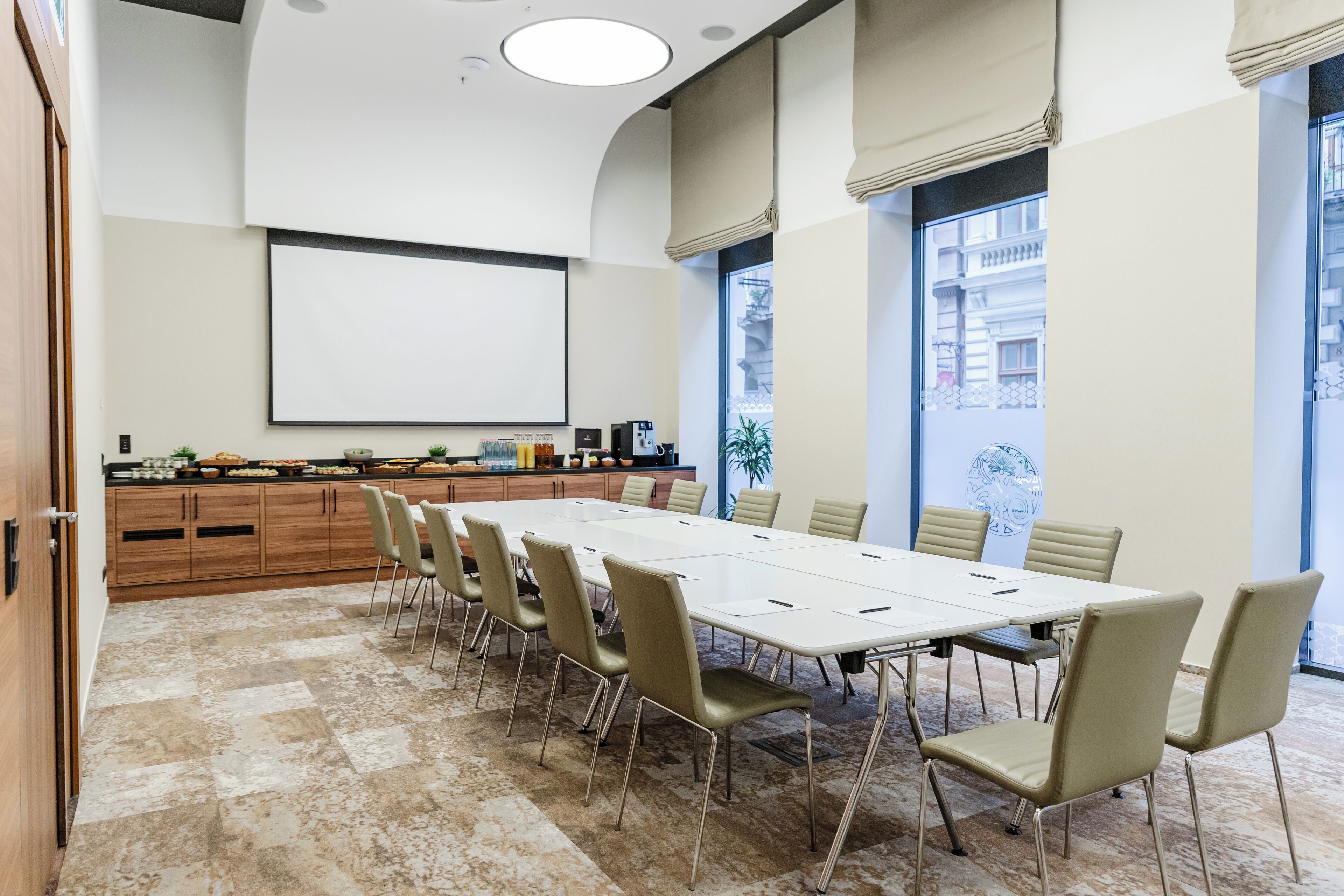 A conference room with a long table, beige chairs, a projector screen, and a large window with a view of buildings.