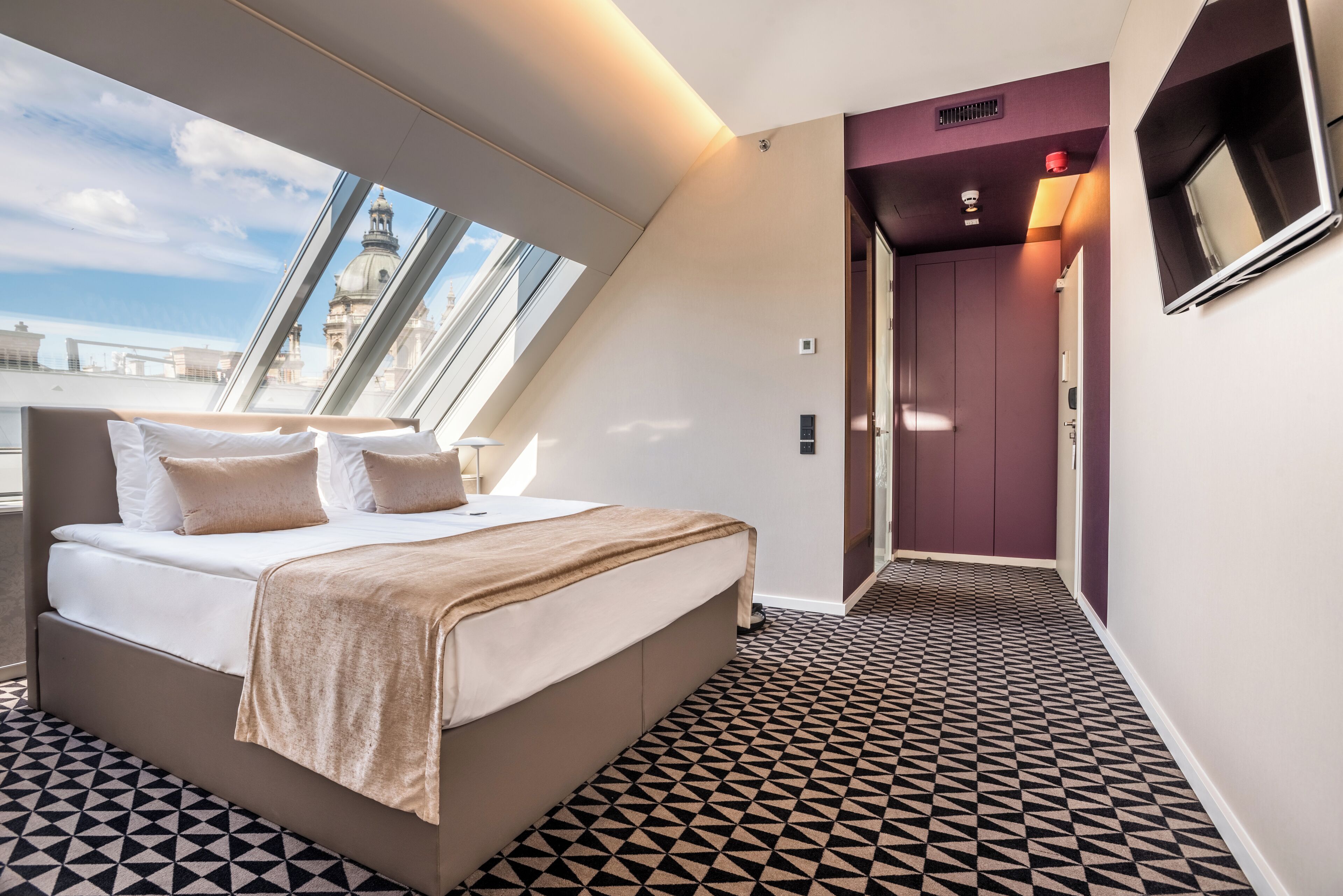 A modern hotel room with a large bed, a view of the cityscape, and a patterned floor.