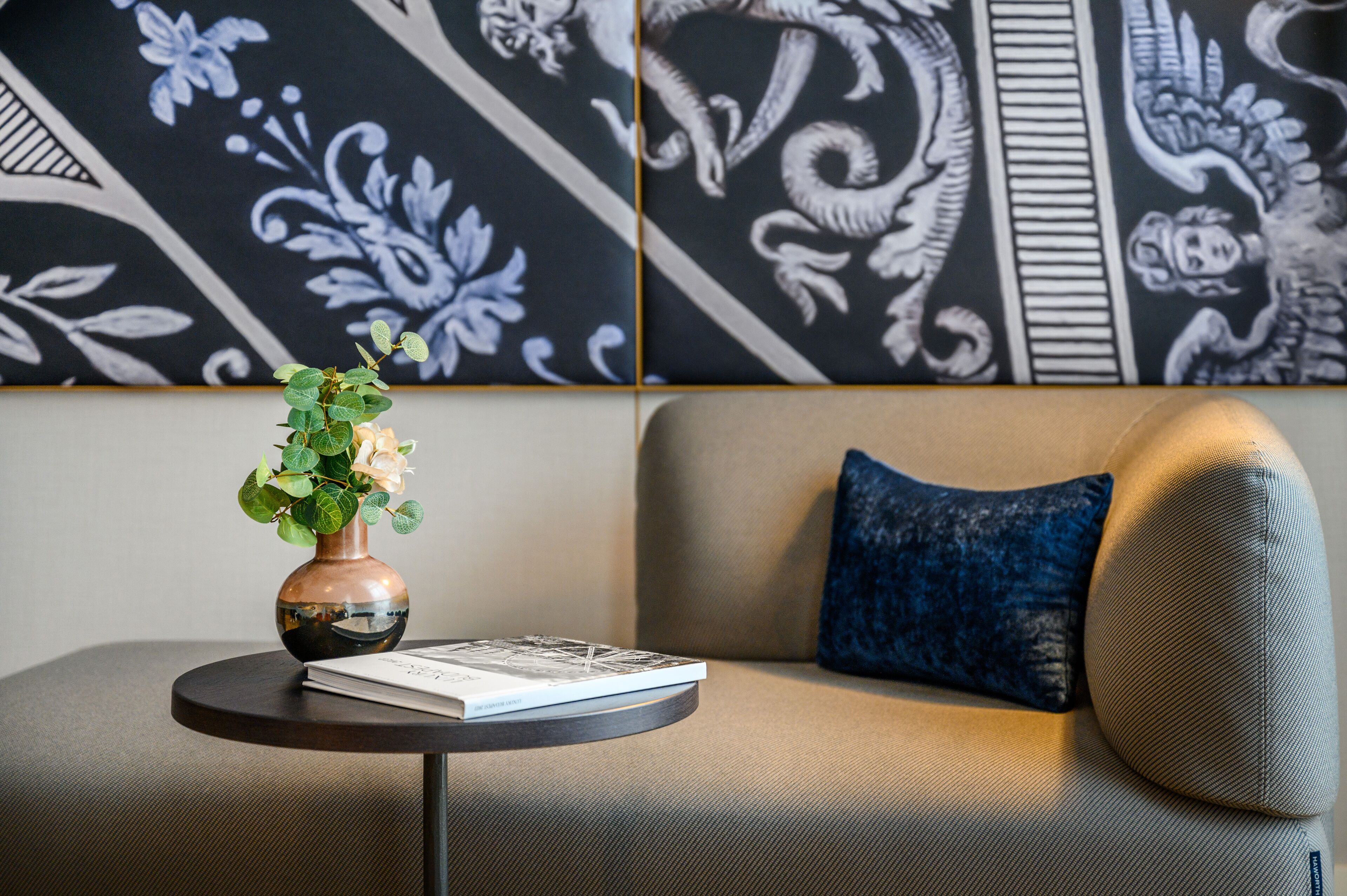 A modern living room with a round table, a vase with flowers, a book, and a sofa with a blue pillow.