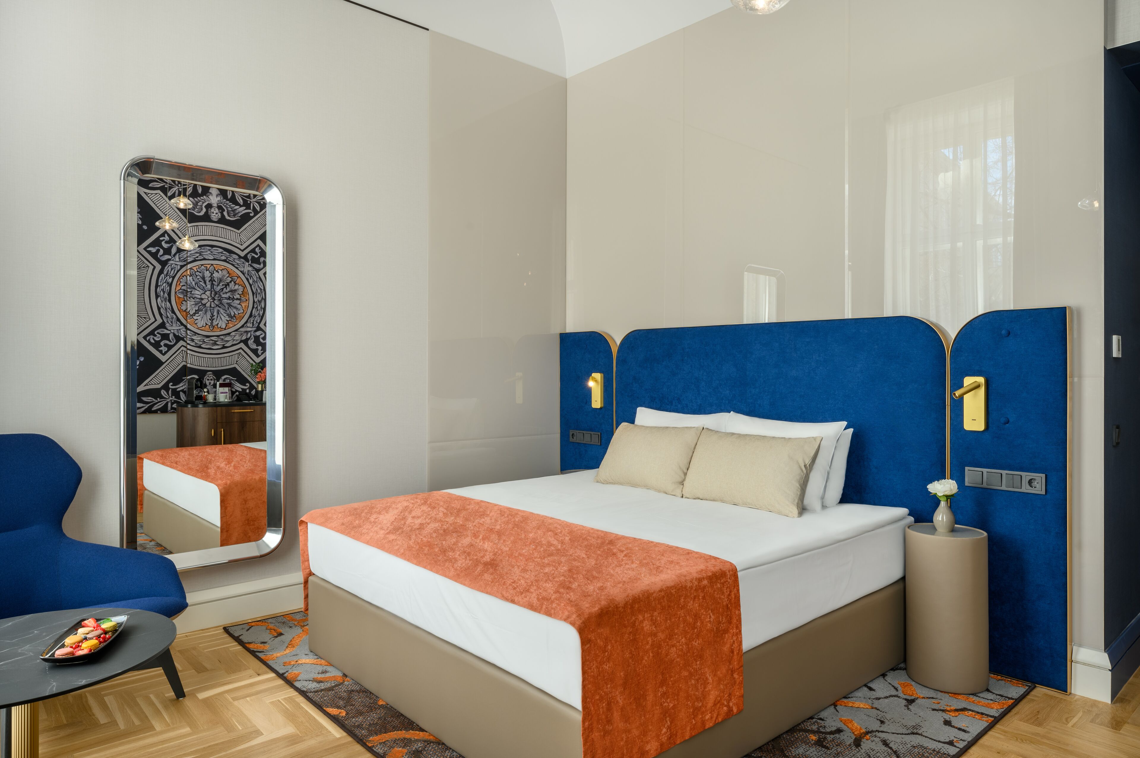Ein modernes Hotelzimmer mit einem großen Bett, einem blauen Sitzmöbel, einem kleinen Tisch und einem Spiegel mit dekorativem Rahmen.