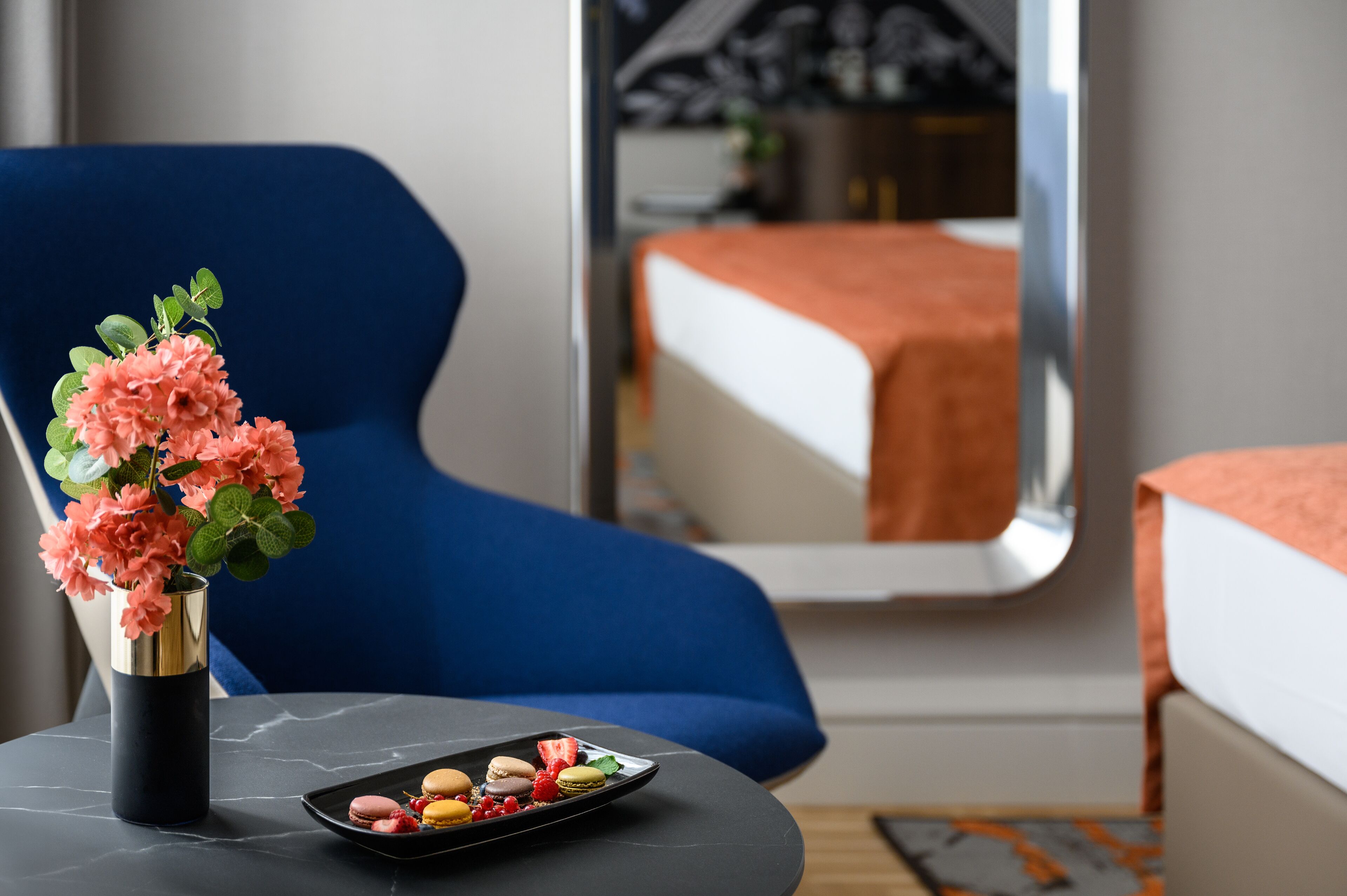 Una habitación de hotel con un sillón azul, una mesita con un florero de flores rosas y un plato de macarons.