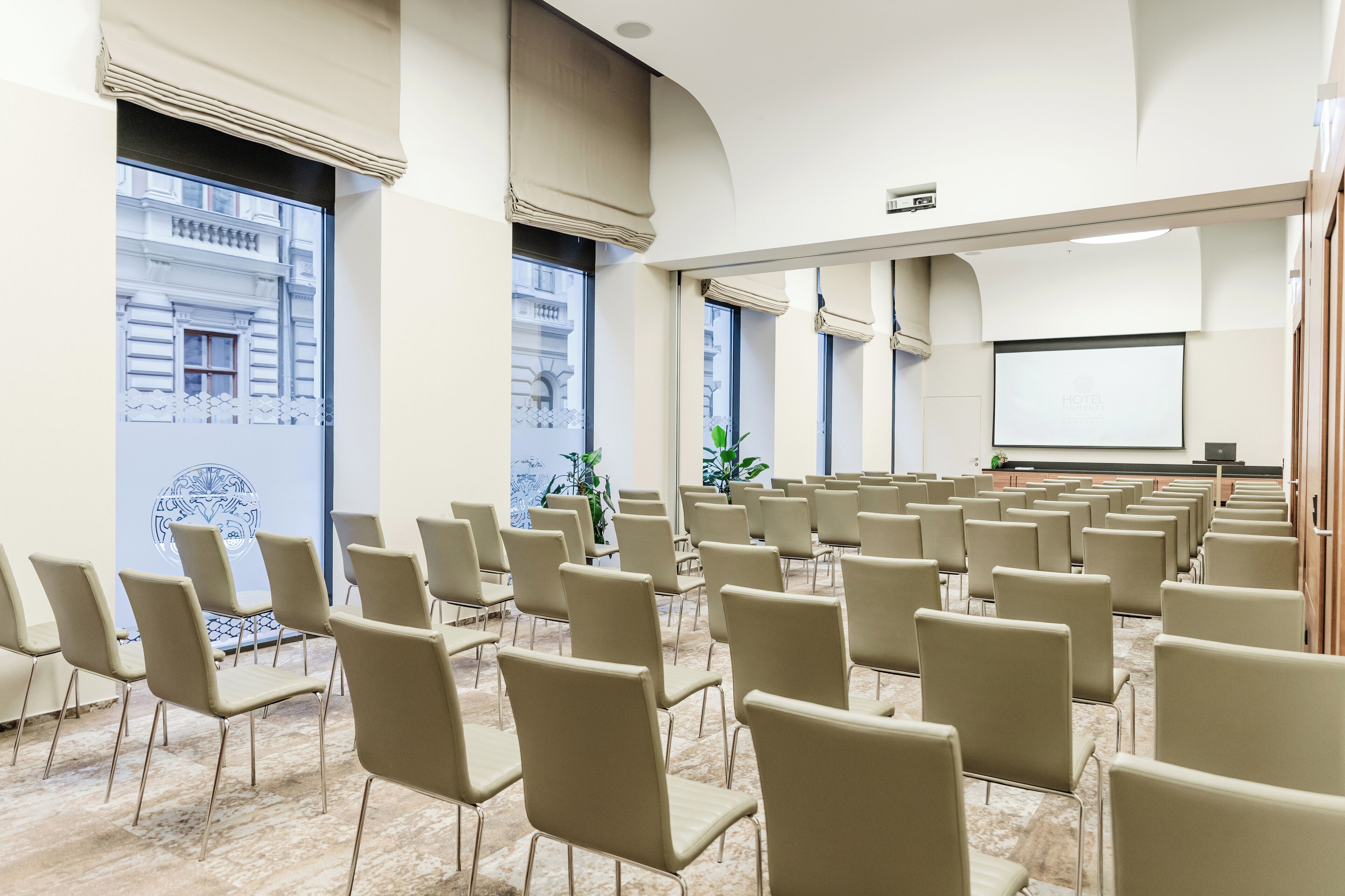 A conference room with rows of chairs, a projector screen, and large windows with views of buildings.