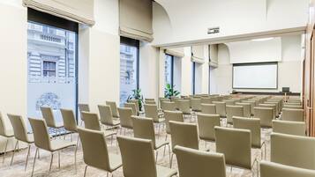 A conference room with rows of chairs, a projector screen, and large windows with views of buildings.