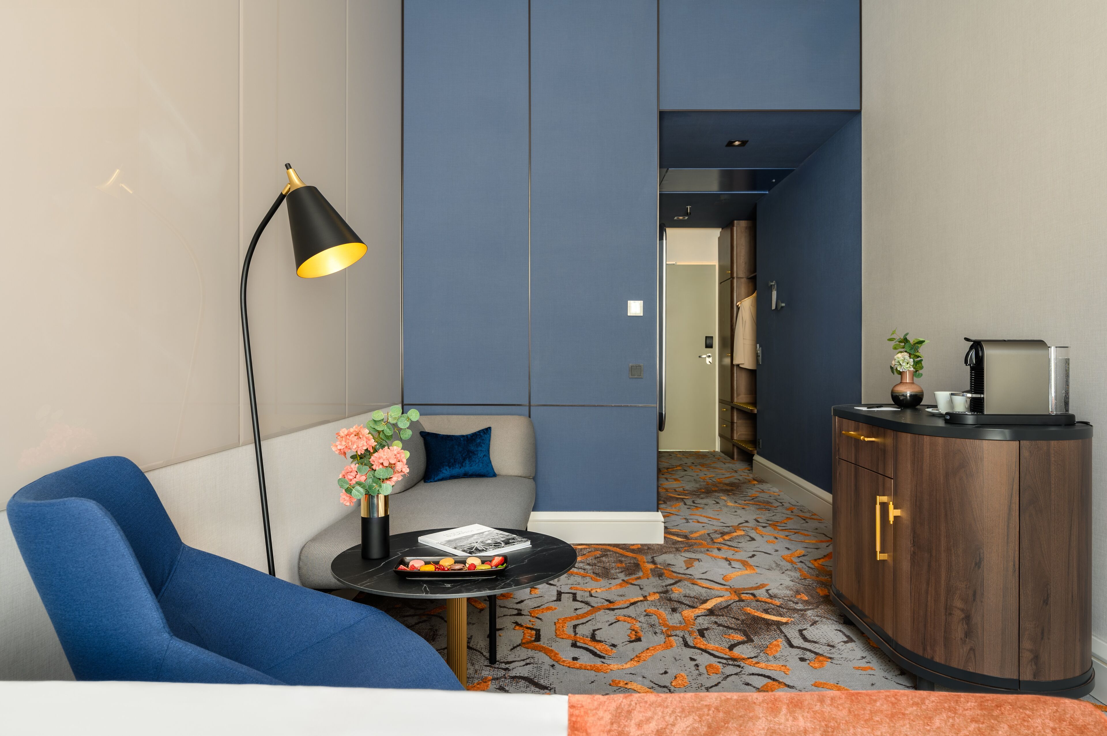 Moderni hotellihuone, jossa on sininen sohva, pieni pyöreä pöytä lampun kanssa, kaappi ja kuviollinen matto.