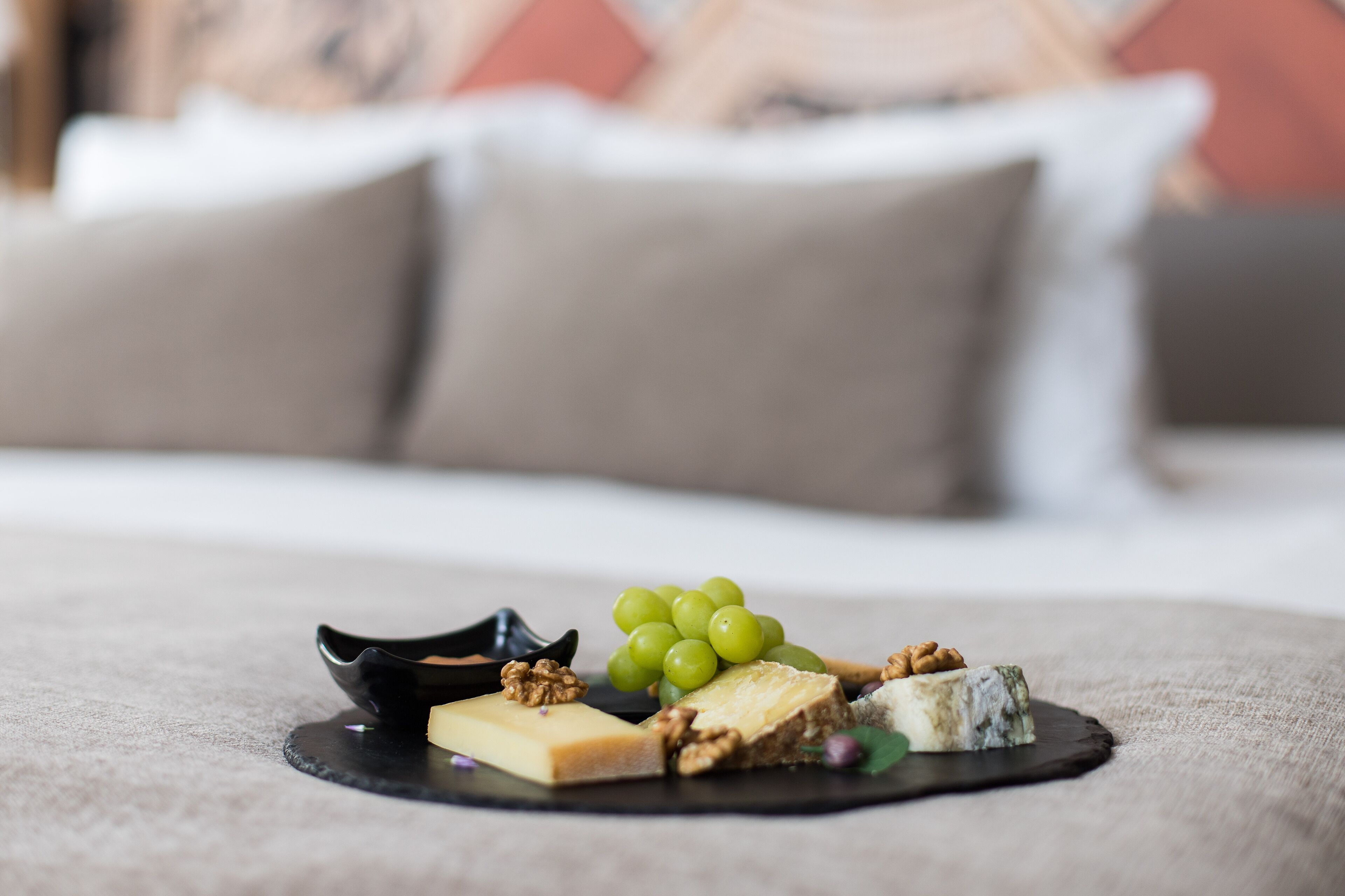 A cheese platter with various cheeses and nuts on a black plate, set on a bed with a beige blanket.