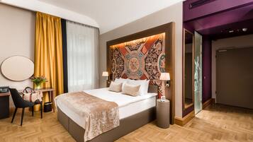 A hotel room with a large bed, a desk with a chair, a small table with a vase, and a mirror on the wall.