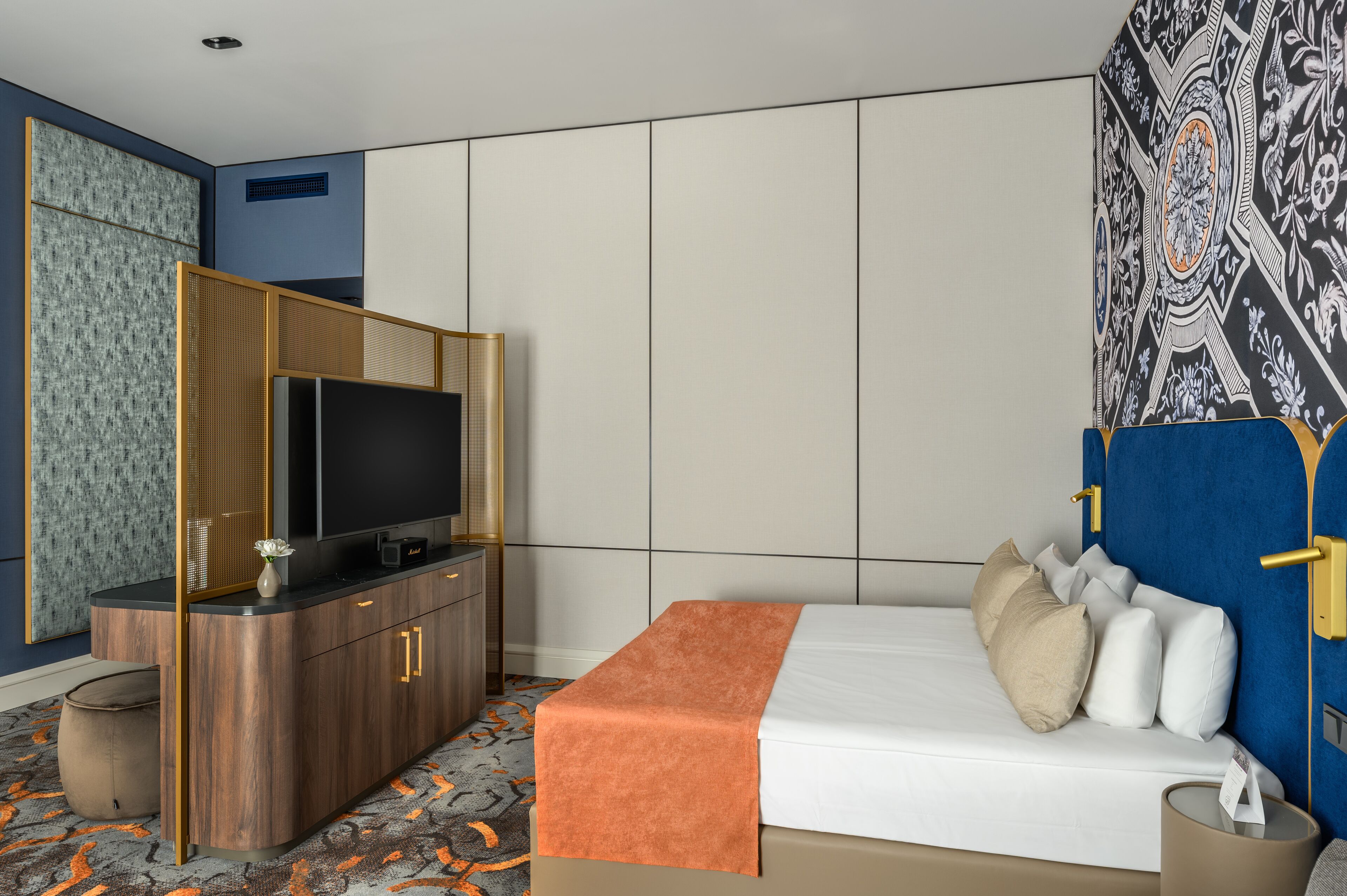 A hotel room with a bed, a TV on a wooden cabinet, a patterned wall, and a blue headboard.