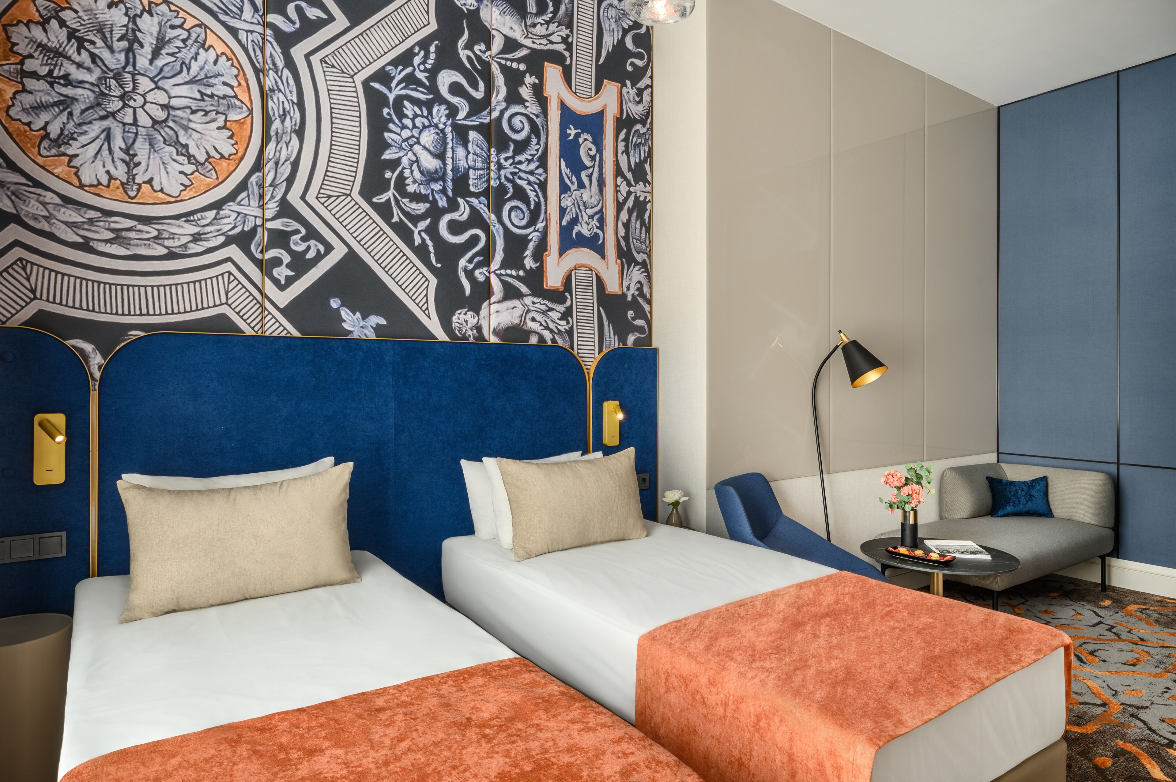 A hotel room with two beds, a sofa, a small table with a lamp, and a decorative wall.