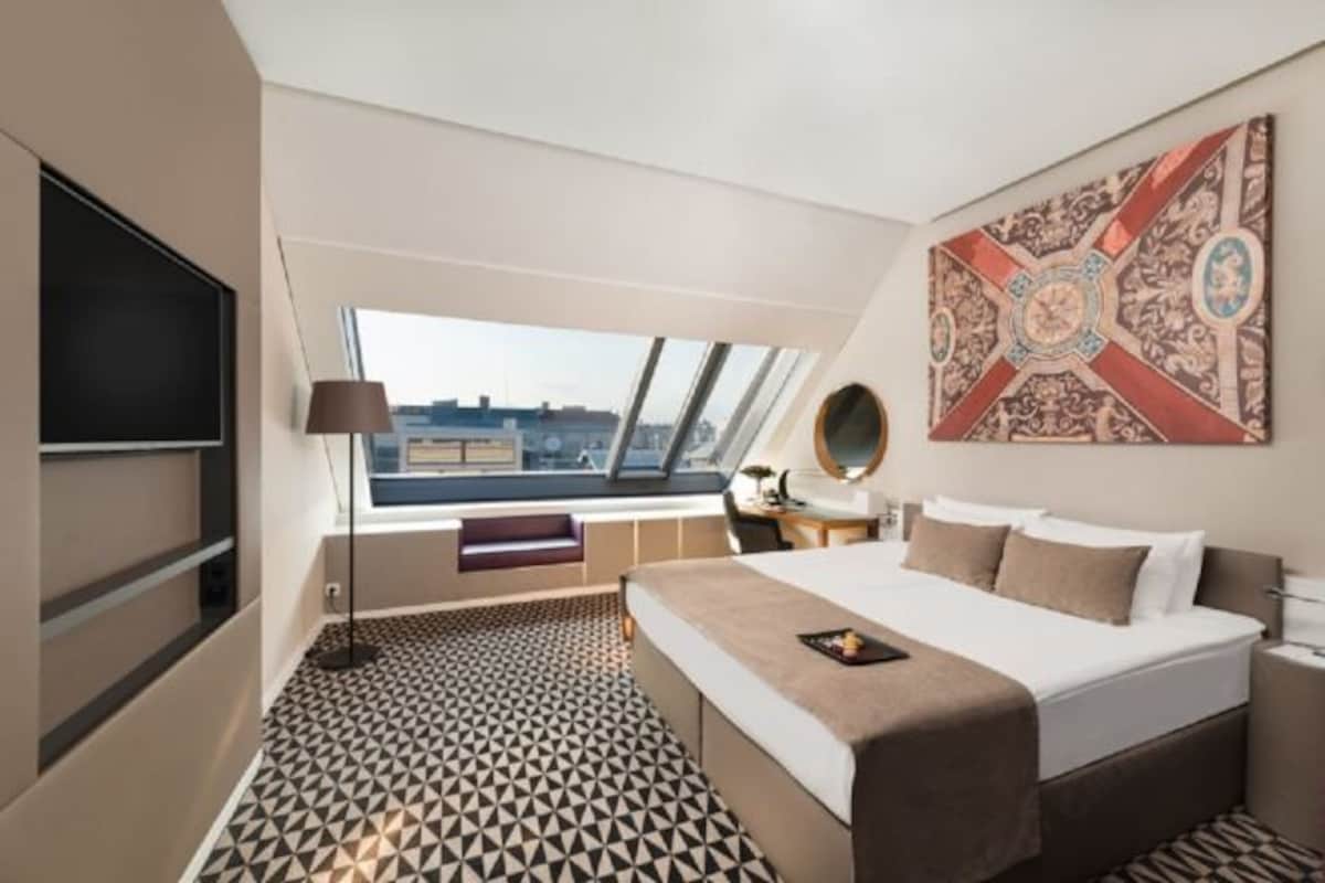 Habitación de hotel con cama, televisión, escritorio, silla, ventana y piso estampado.