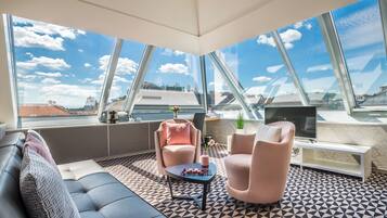 Un área de descanso moderna con piso de diseño geométrico, un sofá, dos sillones, una mesa de centro con un florero y aperitivos, un televisor sobre un mueble bajo y ventanas amplias con vista a la ciudad.