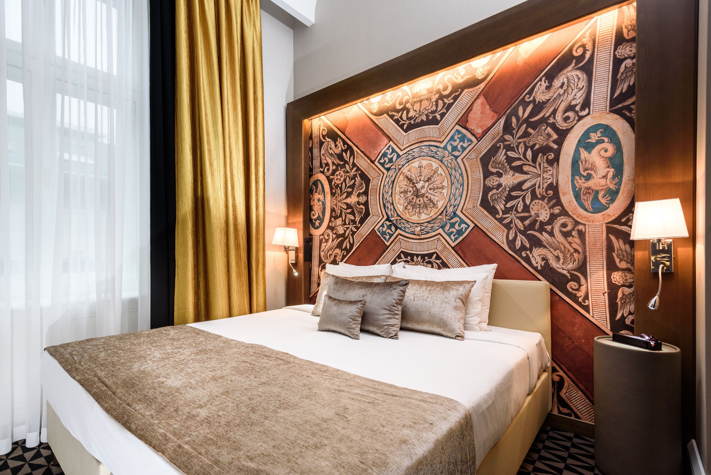 A hotel room with a large bed, a decorative headboard, a nightstand with a lamp, and a window with sheer curtains.