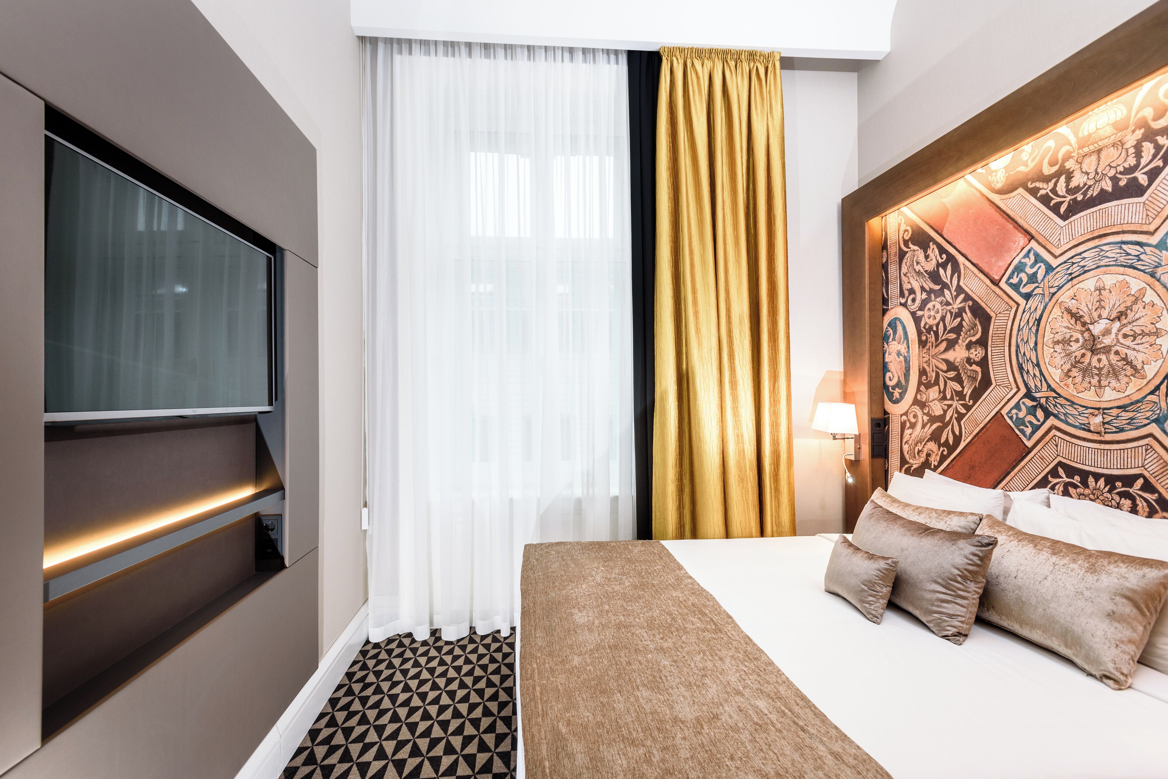 A hotel room with a bed, a TV, a patterned headboard, a large window with sheer curtains, and a patterned floor.