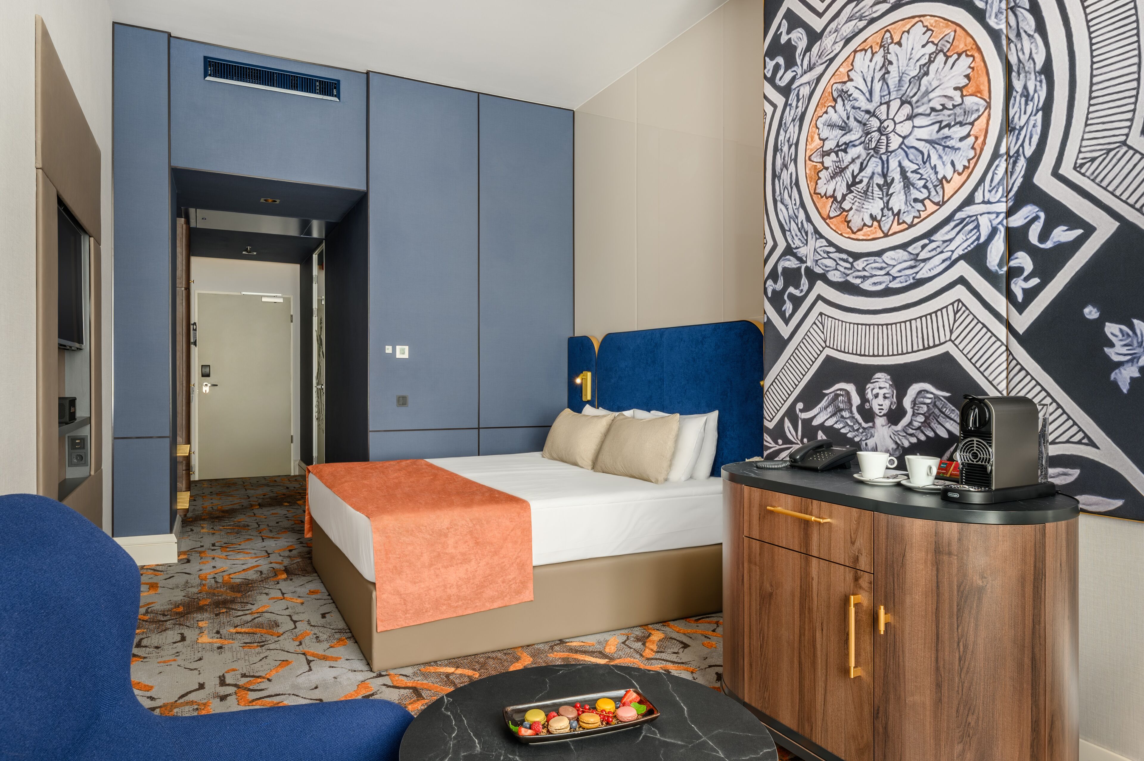 A hotel room with a bed, a desk with a coffee maker, a tray of snacks, and a decorative wall.