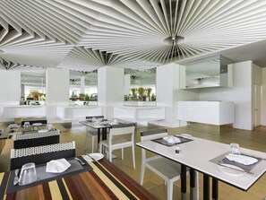 Um saguão de hotel moderno com design de teto singular, paredes espelhadas e área de refeições com mobiliário branco.