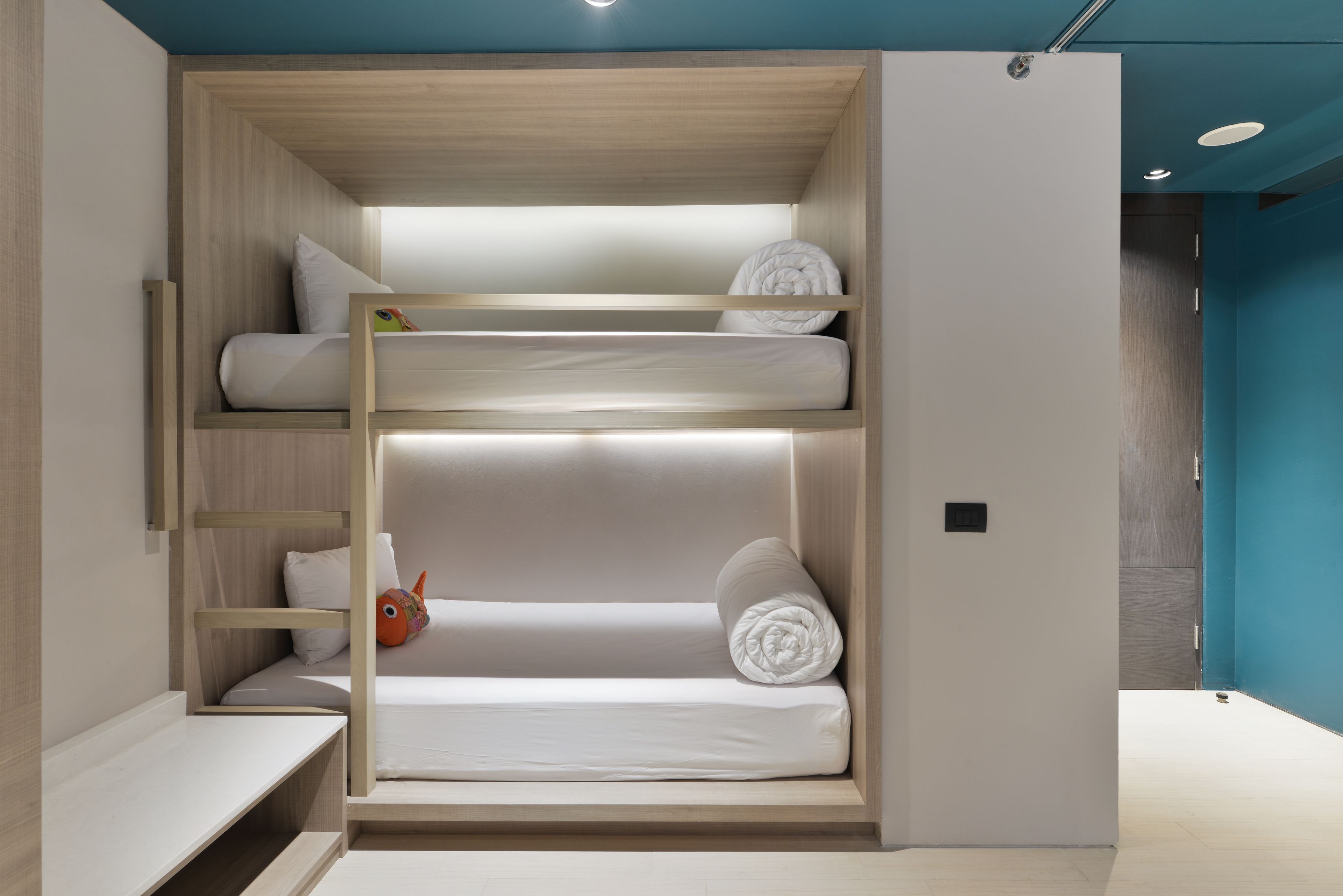 Ein Stockbett mit Holzrahmen, weißer Bettwäsche und einem kleinen Regal mit einem Spielzeug.
