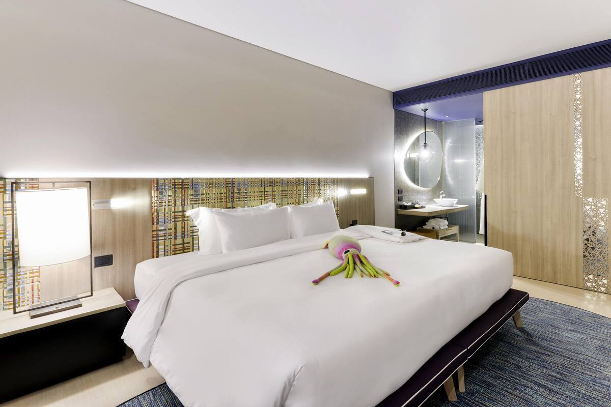 Moderni hotellihuone, jossa on suuri sänky, yöpöytä, peili ja kylpyhuone, joka on näkyvissä väliseinän läpi.