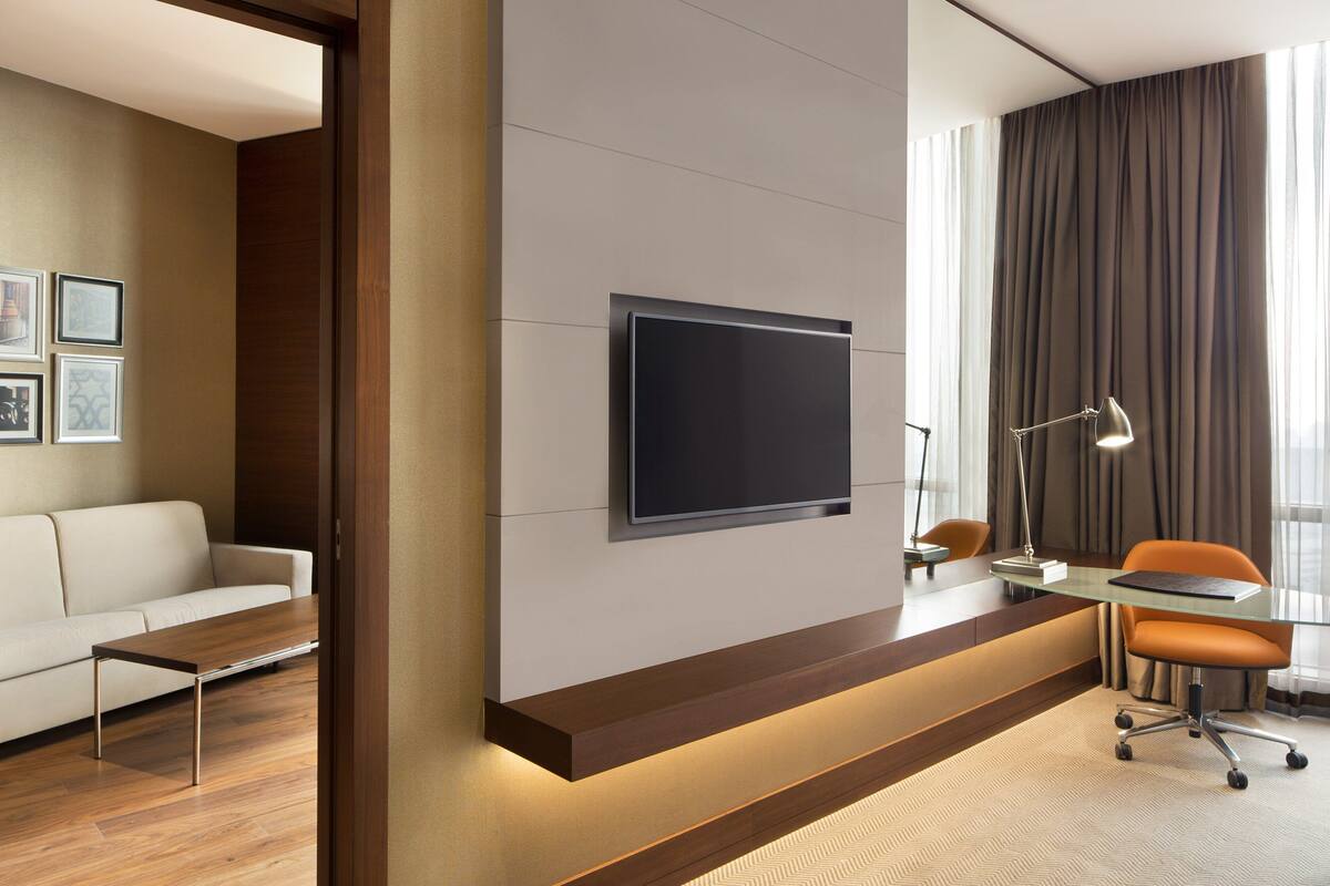 Ein modernes Hotelzimmer mit einem an der Wand befestigten Flachbildfernseher, einem Holztischchen, einer Couch, einem orangefarbenen Sessel und einem Schreibtisch mit Lampe.
