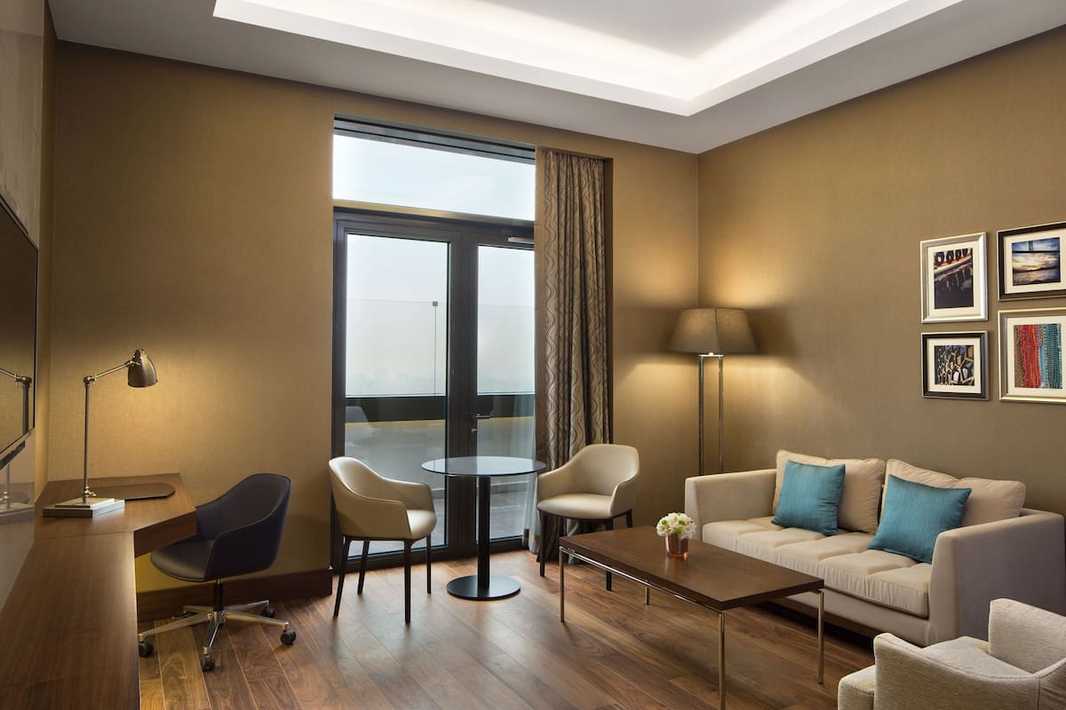 Ein modernes Hotelzimmer mit Holzboden, einer Couch, zwei Sesseln, einem Couchtisch, einem Schreibtisch mit Lampe und einem großen Fenster mit Vorhängen.
