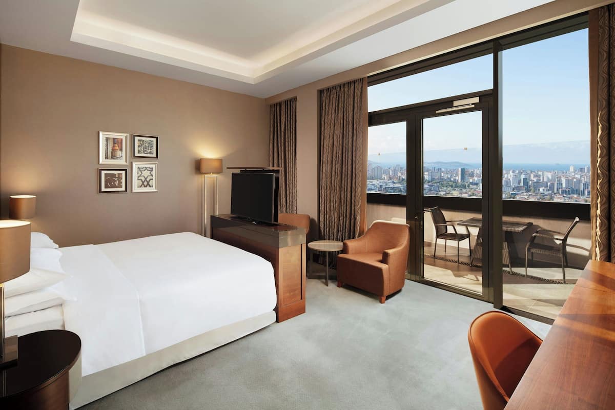 Ein Hotelzimmer mit einem großen Bett, einem Schreibtisch mit Fernseher, einem kleinen runden Tisch, einem Sessel und einem Balkon mit Blick auf die Stadt.