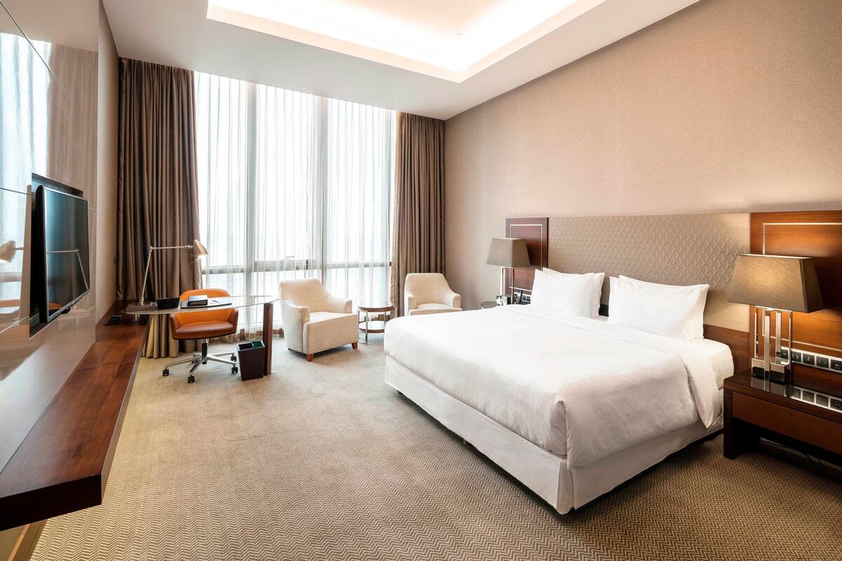 Ein Hotelzimmer mit einem großen Bett, zwei Sesseln, einem Schreibtisch mit Stuhl, einem Fernseher und einem großen Fenster mit Vorhängen.