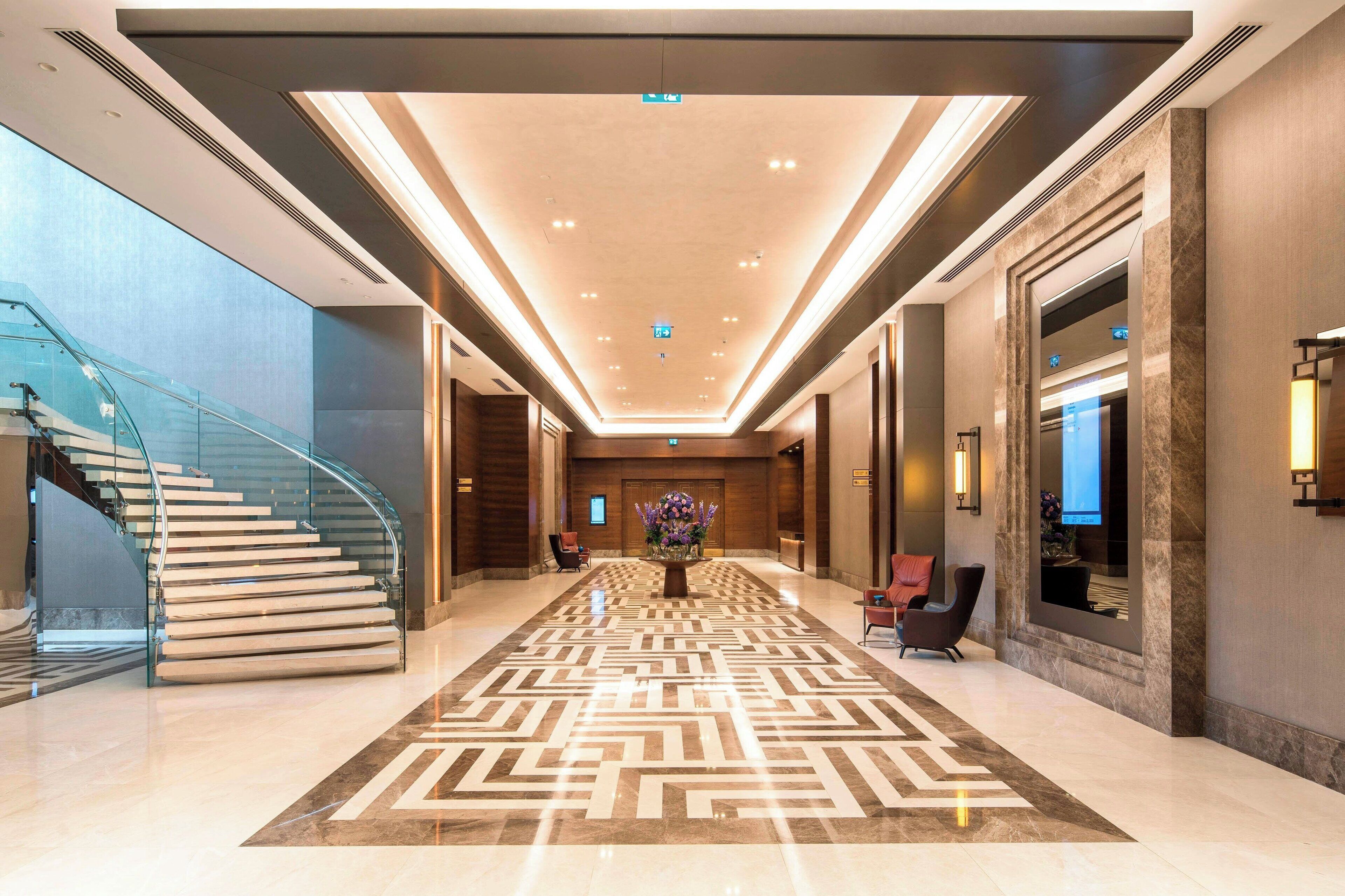 A modern hotel lobby with a glass staircase, a patterned carpet, and a central floral arrangement.