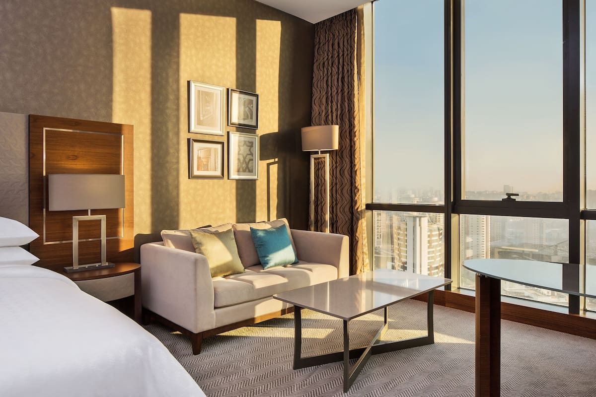 Ein modernes Hotelzimmer mit einem großen Bett, einer Couch, einem Couchtisch und Blick auf die Stadt durch große Fenster.