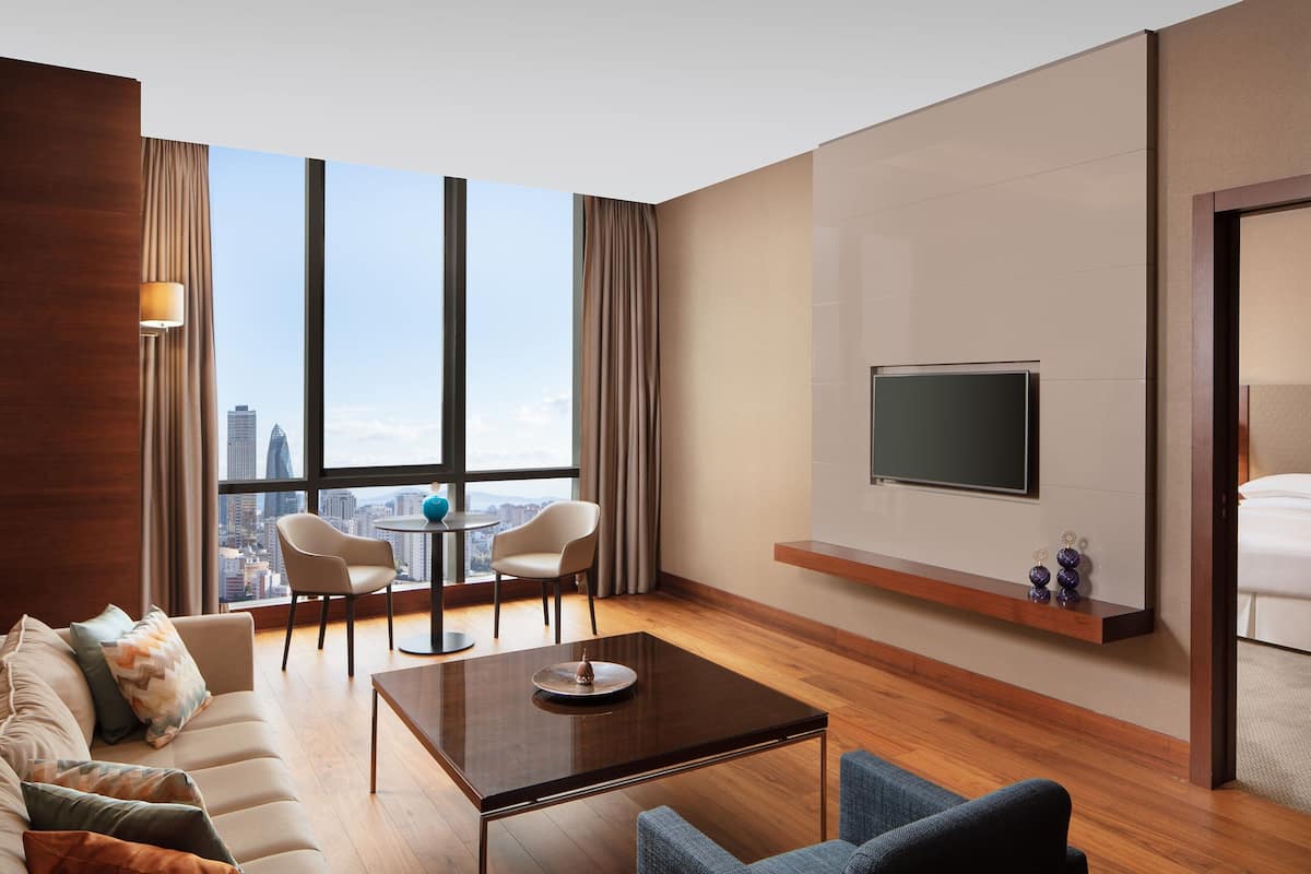 Ein modernes Hotelzimmer mit großem Fenster und Blick auf die Stadt, einem flachbildfernseher an der Wand, einem Sofa, einem Couchtisch und einer Essecke mit Stühlen.