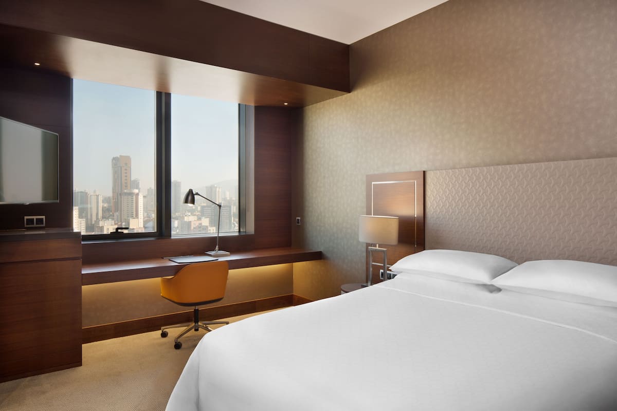 Ein modernes Hotelzimmer mit einem großen Bett, einem Schreibtisch mit Stuhl, einem Fenster mit Stadtblick und einem Nachttisch mit Lampe.