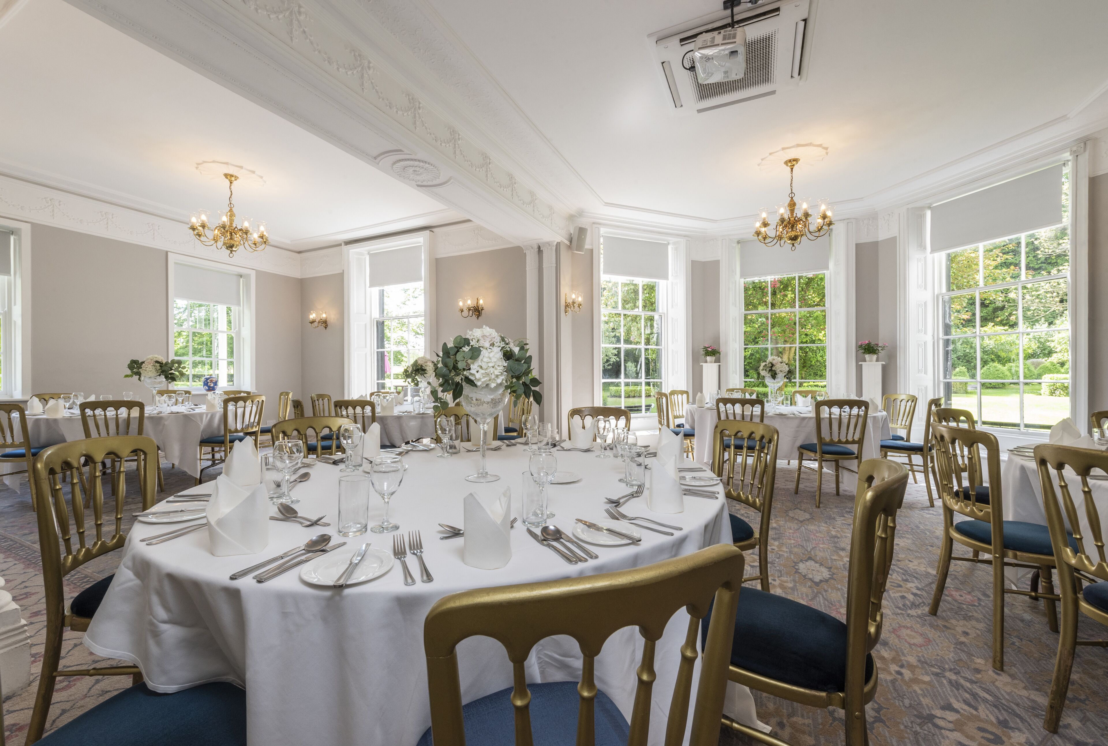 A formal dining room with round tables, gold chairs, and chandeliers.