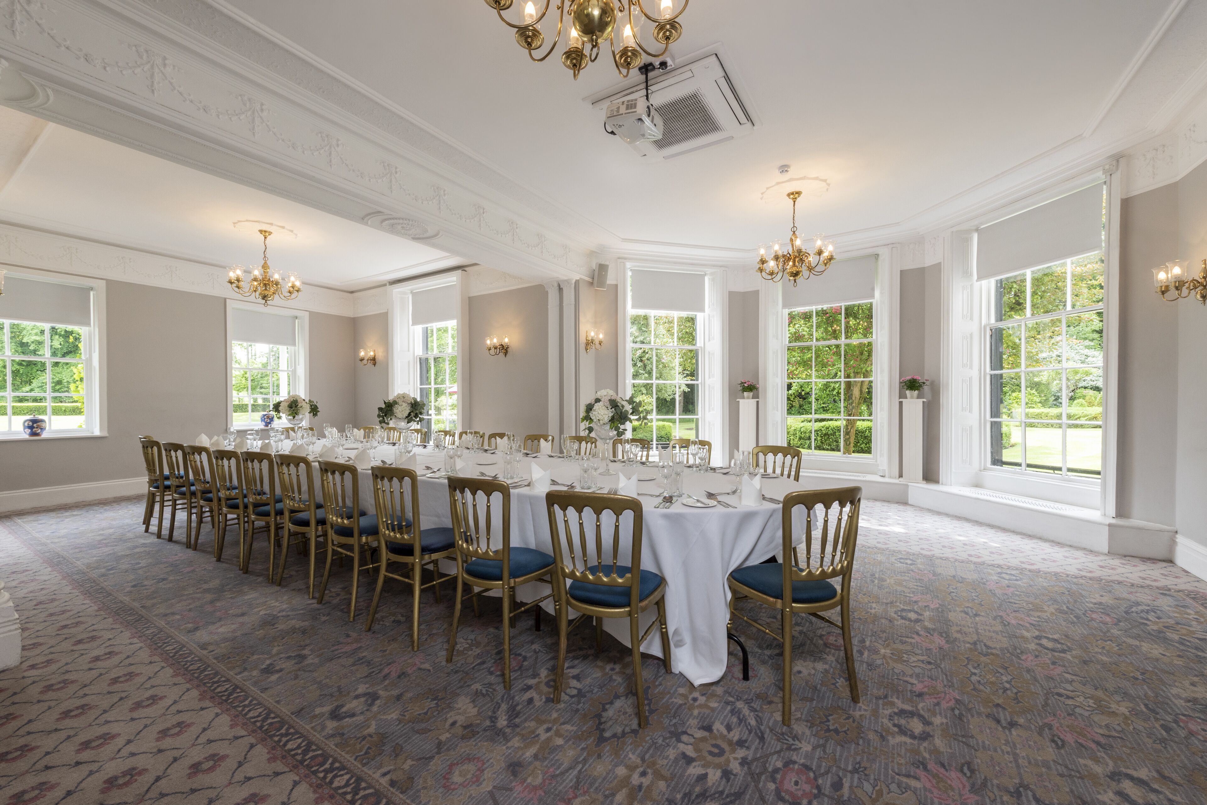 A formal dining room with a long table set for a meal, surrounded by chairs with blue cushions.