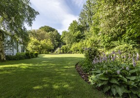 A well-maintained garden with a lawn, a white building with black trim, and dense foliage.