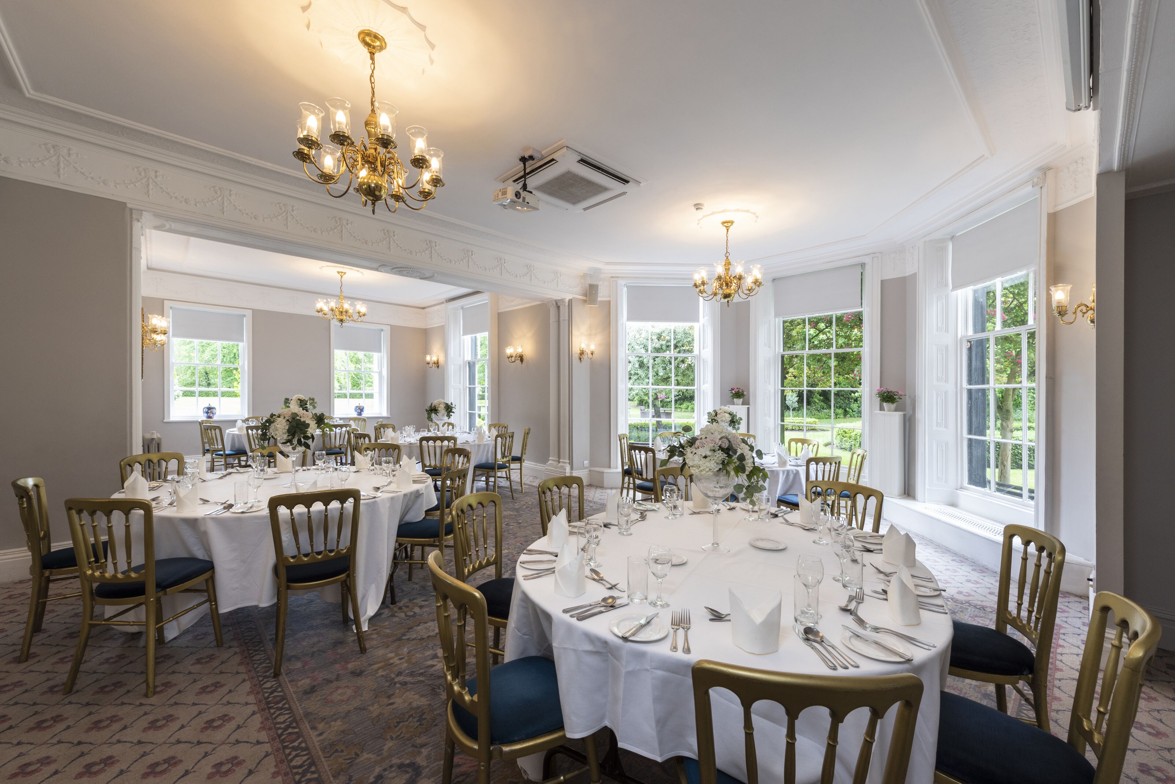 A formal dining room with round tables, gold chairs, and white tablecloths.
