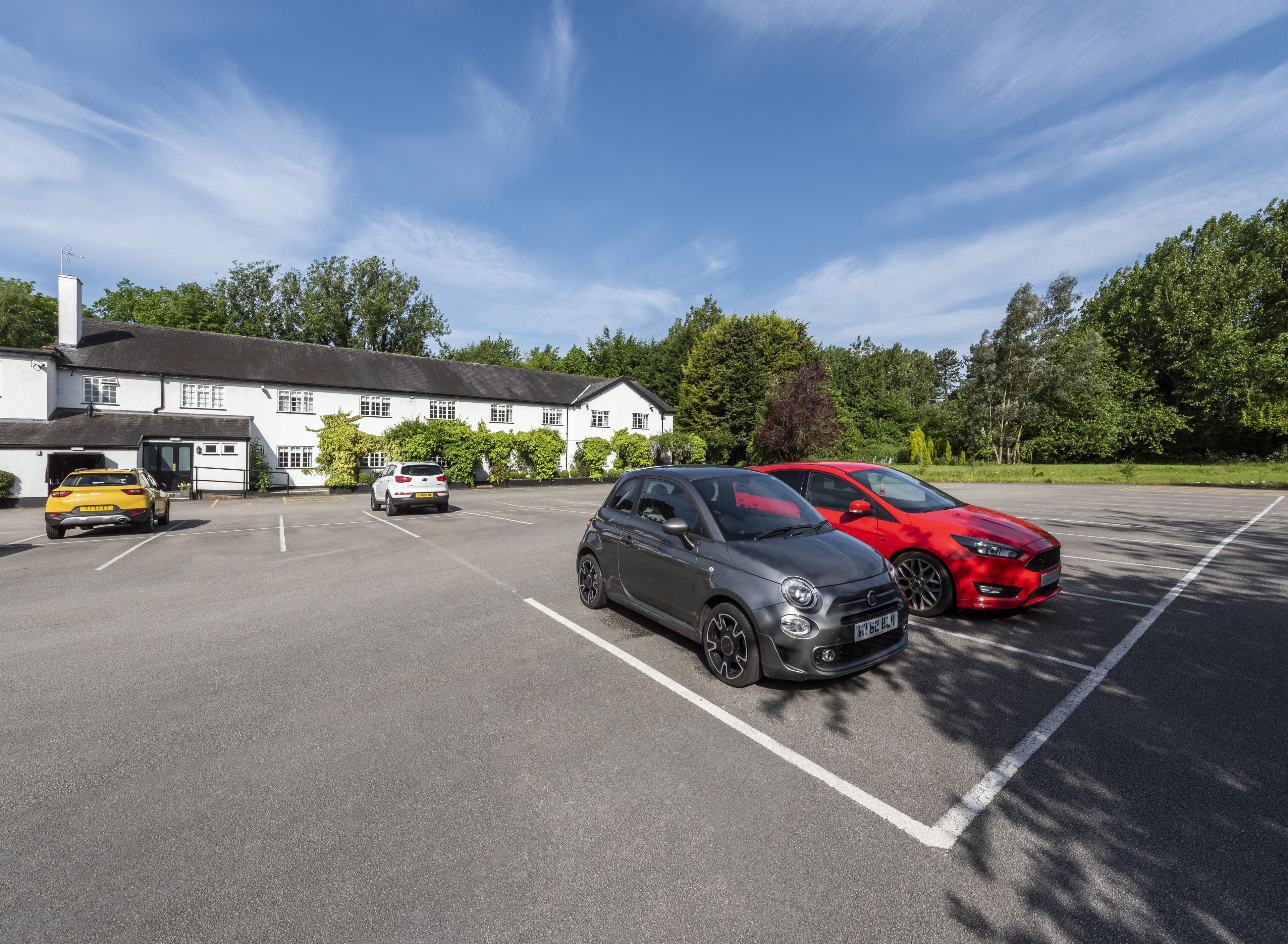 A parking lot with several cars, a white building with a black roof, and trees in the background.