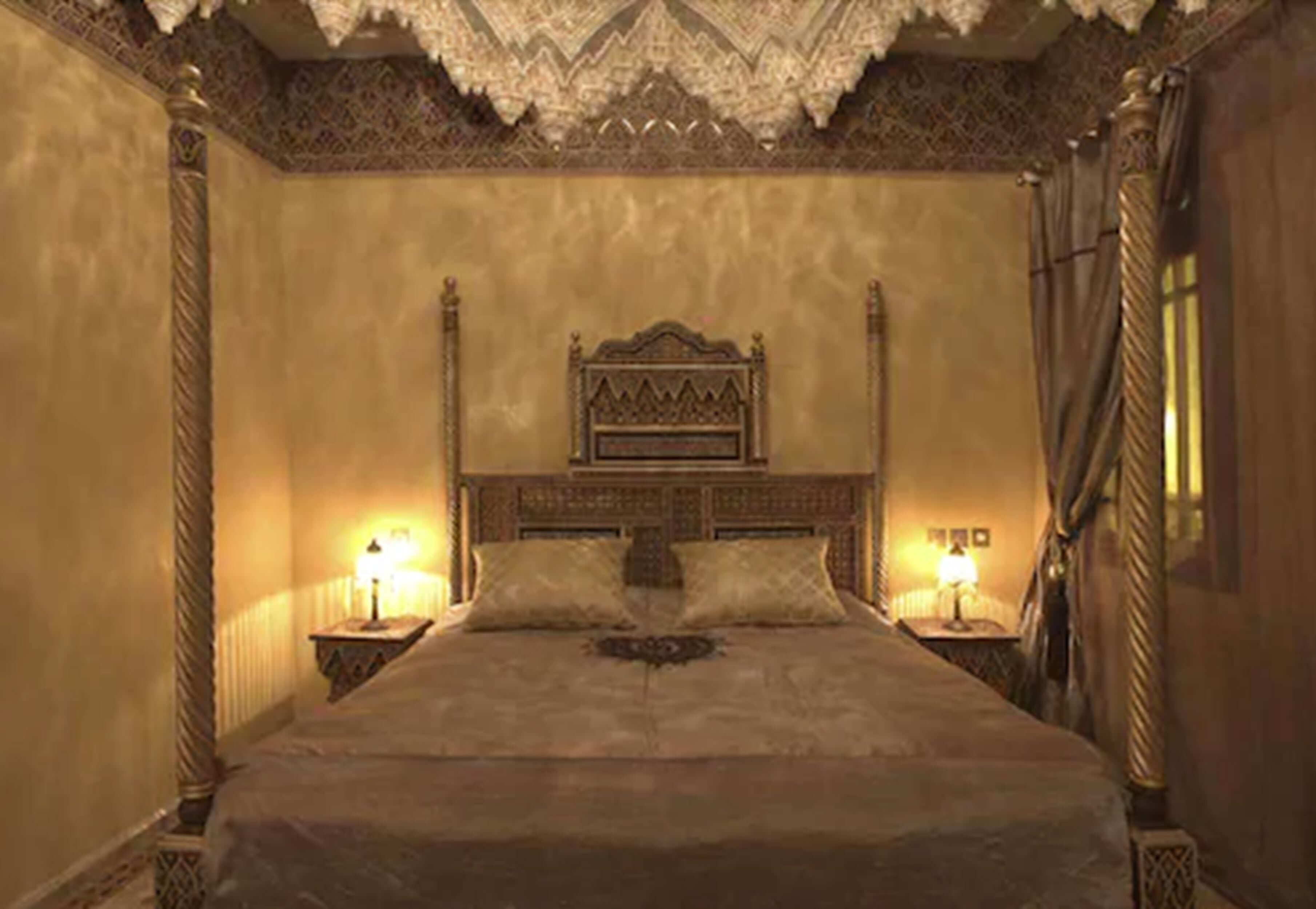 A large, ornate bed with a high headboard, flanked by two bedside tables with lamps, in a room with patterned walls and a decorative ceiling.