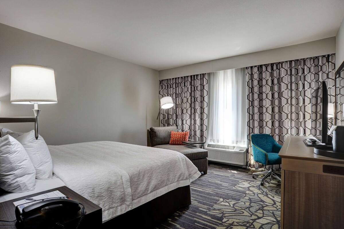 A hotel room with a bed, bedside lamp, desk, chair, and a window with patterned curtains.