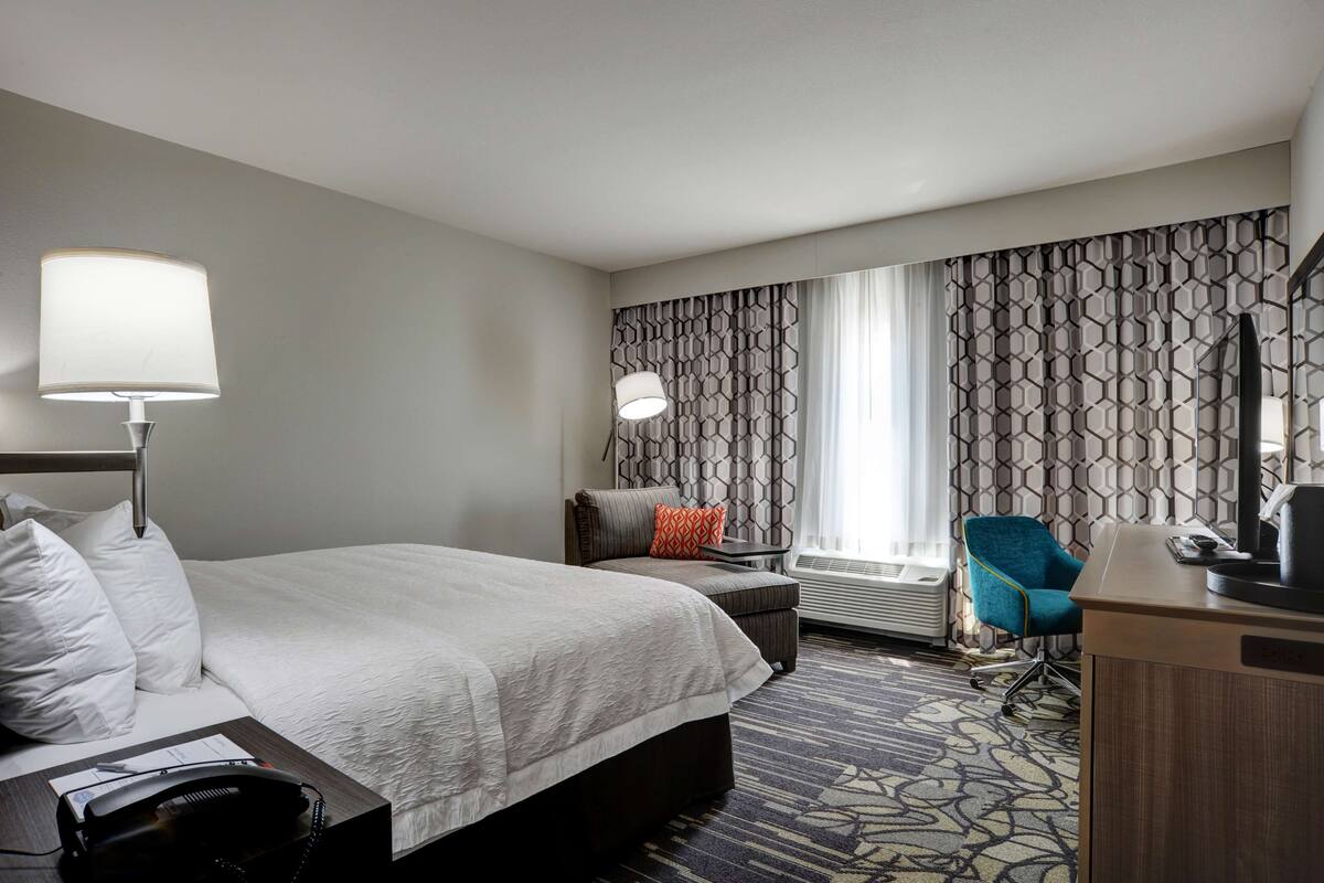 A hotel room with a bed, desk, chair, lamp, and patterned curtains.