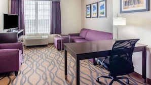 A hotel room with a sofa, a desk, a chair, a TV, and a window with curtains. - La Quinta Inn & Suites by Wyndham Luling (Luling)