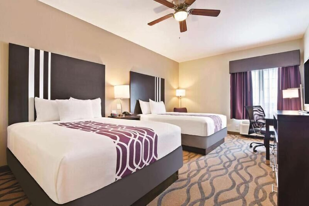 A hotel room with two beds, a desk, a chair, a ceiling fan, and a window with curtains.