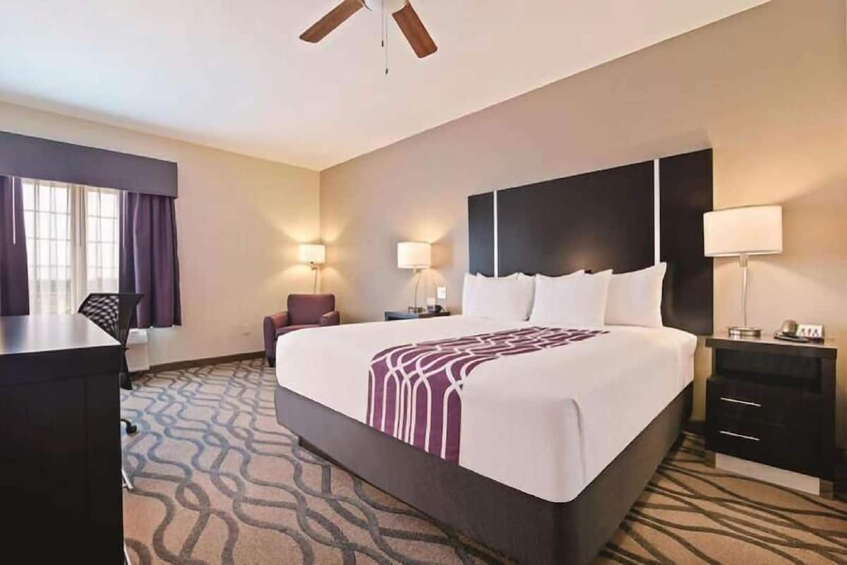 A hotel room with a large bed, a desk, two bedside tables, a ceiling fan, and a window with curtains.