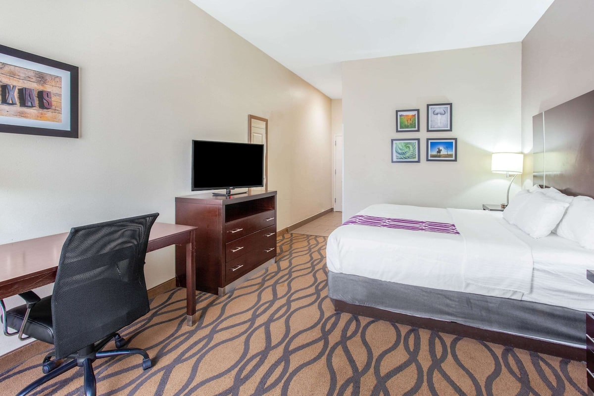 A hotel room with a bed, a desk with a chair, a television, a dresser, and framed pictures on the wall.