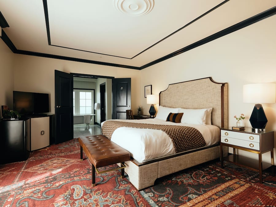 A hotel room with a large bed, a bench, a bedside table with a lamp, a TV, and a wardrobe.
